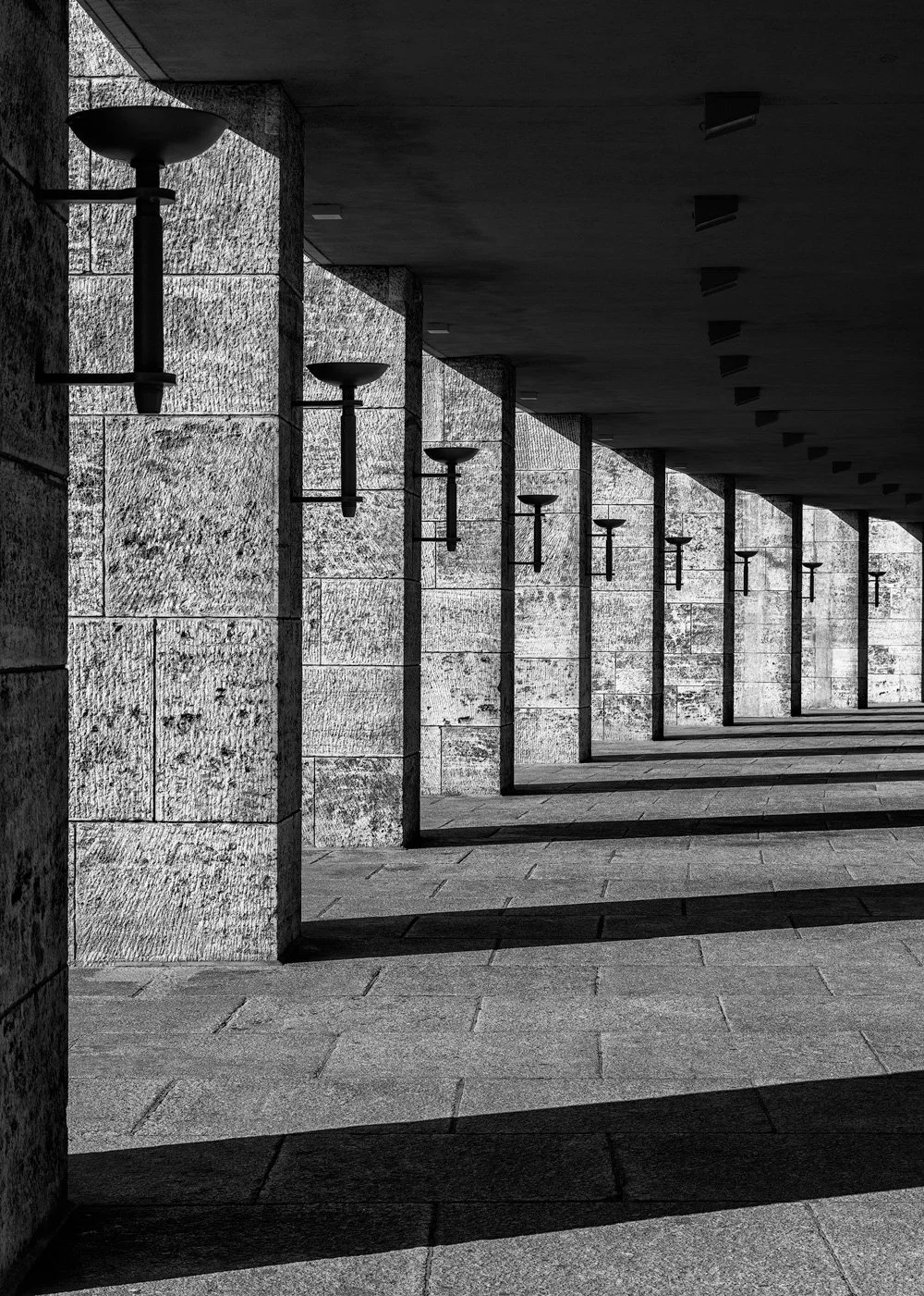 [architecture] [europe] [Berlin] [blackandwhite] [abstract] A receding stone colonnade lined with lantern-topped columns stretches along Berlin's Olympiastadion in monochrome. Architectural Photography by Jim Scott