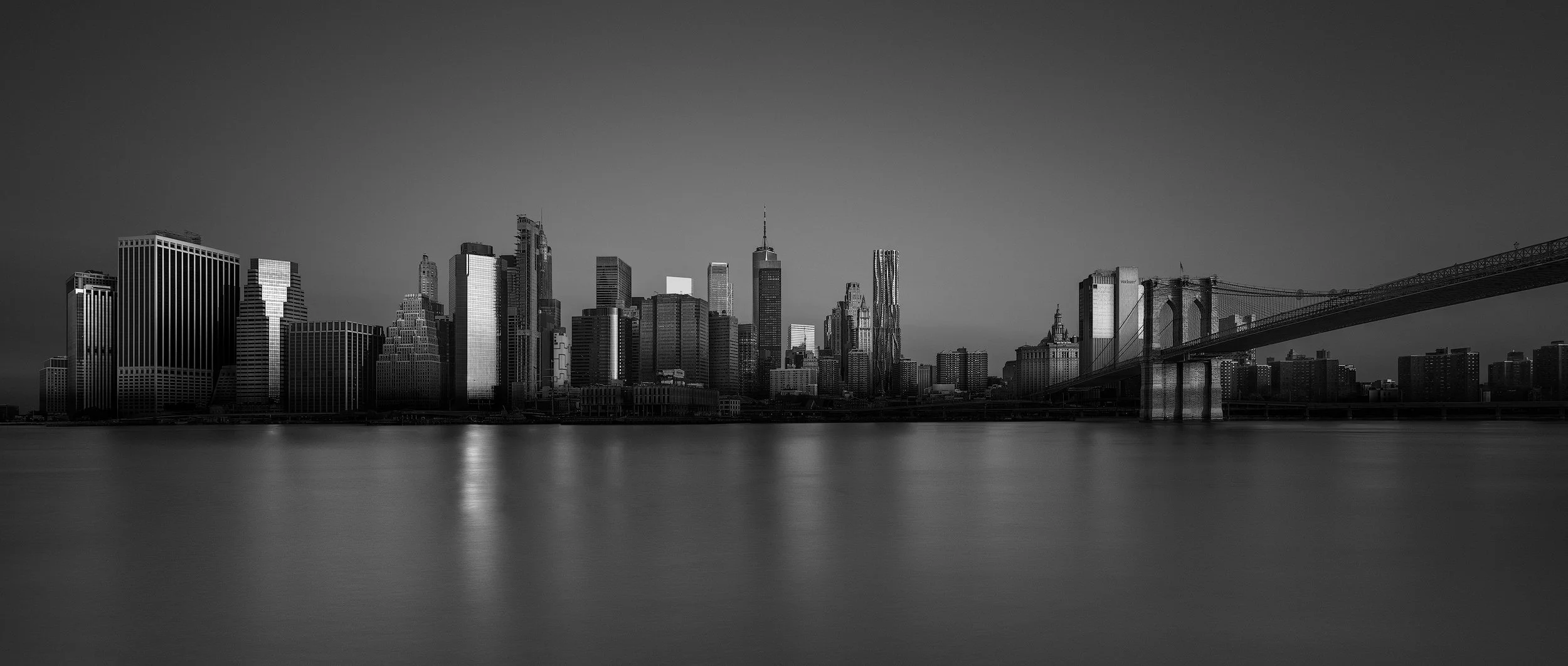 Contemporary and historic commercial architecture in Manhattan, New York: A dramatic monochromatic study of the Lower Manhattan skyline and the Brooklyn Bridge. This panoramic architectural photograph captures the dense cluster of financial district 