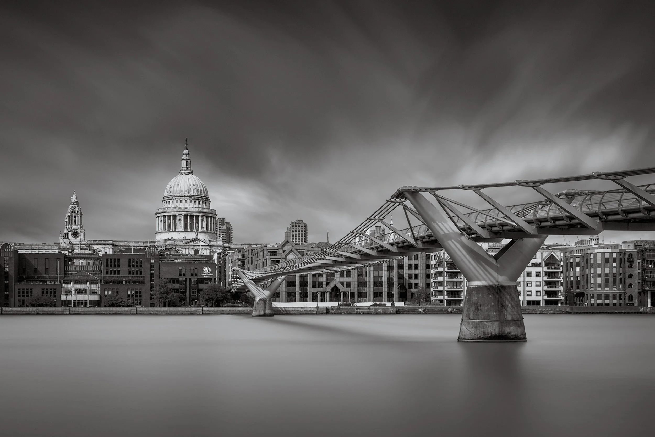 Iconic London architecture: The Millennium Bridge leading to St Paul's Cathedral. A wide-angle black and white long-exposure photograph across the River Thames, showcasing the modern steel 'blade' design of the pedestrian bridge contrasted against Si