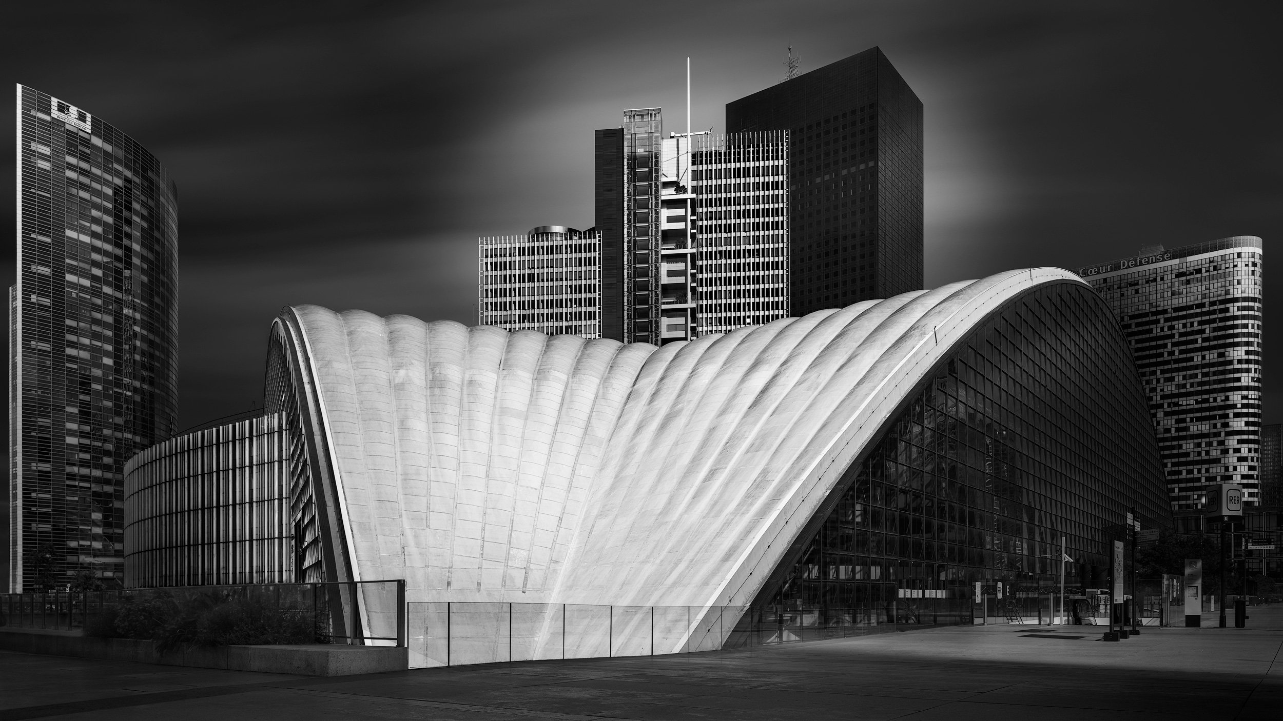 [architecture] [europe] [paris] [blackandwhite] [panoramic] CNIT La Défense Paris B&W, vast ribbed concrete shell vault arching low over the plaza with dark office towers beyond. Architectural Photography by Jim Scott.
