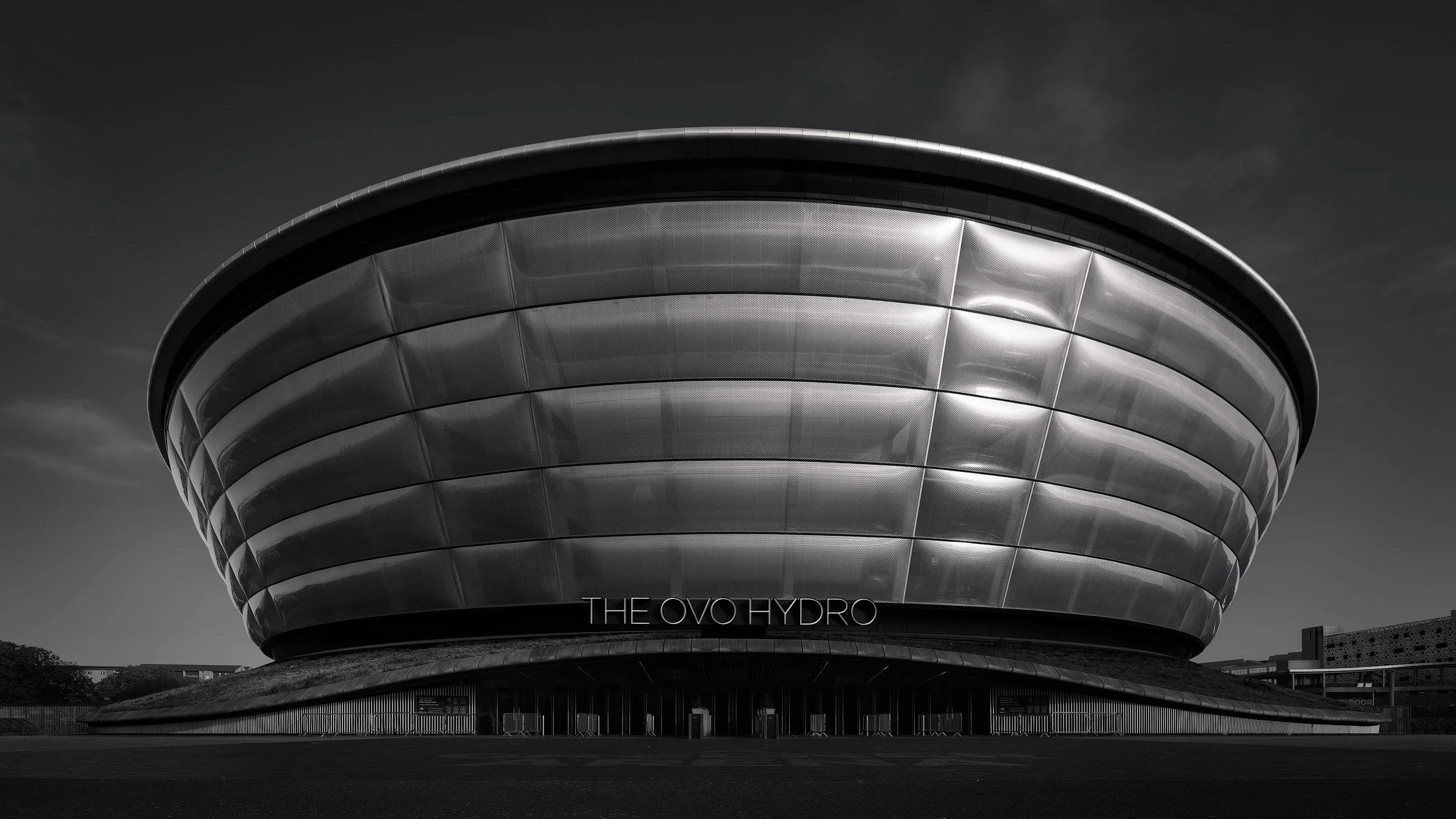 Contemporary arena architecture in Glasgow, Scotland: A symmetrical monochromatic study of the OVO Hydro, designed by Foster + Partners. This architectural photograph captures the rhythmic quilted texture of the ETFE pillowed facade and the building'