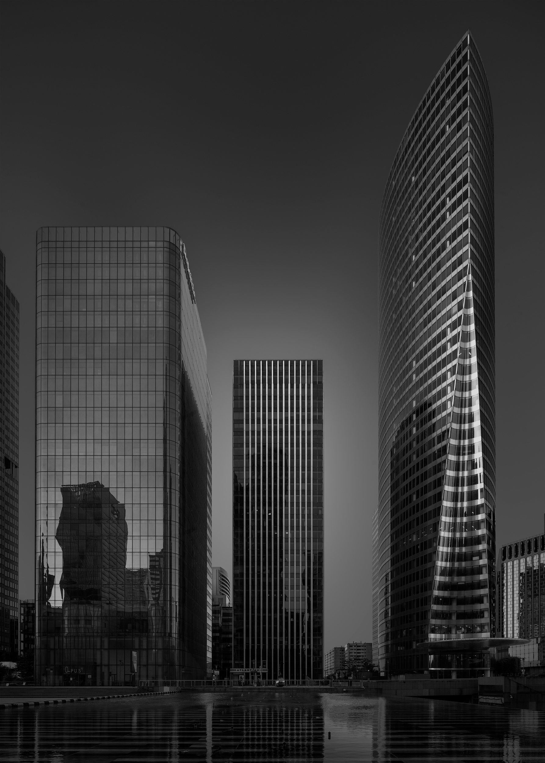 [architecture] [europe] [paris] [blackandwhite] La Defense Paris esplanade, trio of contrasting glass office towers reflected in water, dark dramatic black and white. Architectural Photography by Jim Scott.