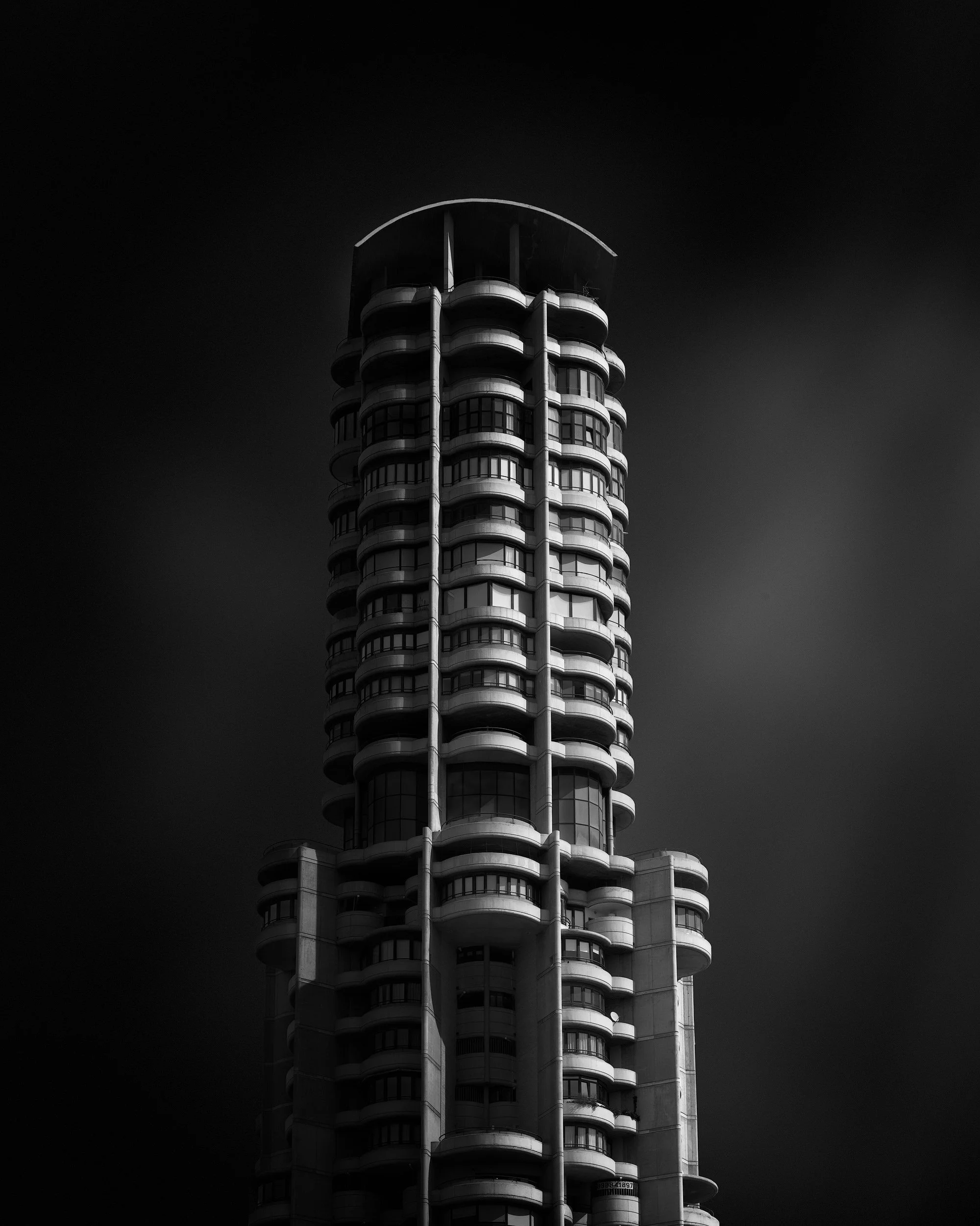 Brutalist residential architecture in Benidorm, Spain: A dramatic monochromatic study of the Torre Lúgano, designed by Adolfo Rodríguez. This architectural photograph captures the tower’s complex, tiered silhouette and rhythmic curved balconies. The 