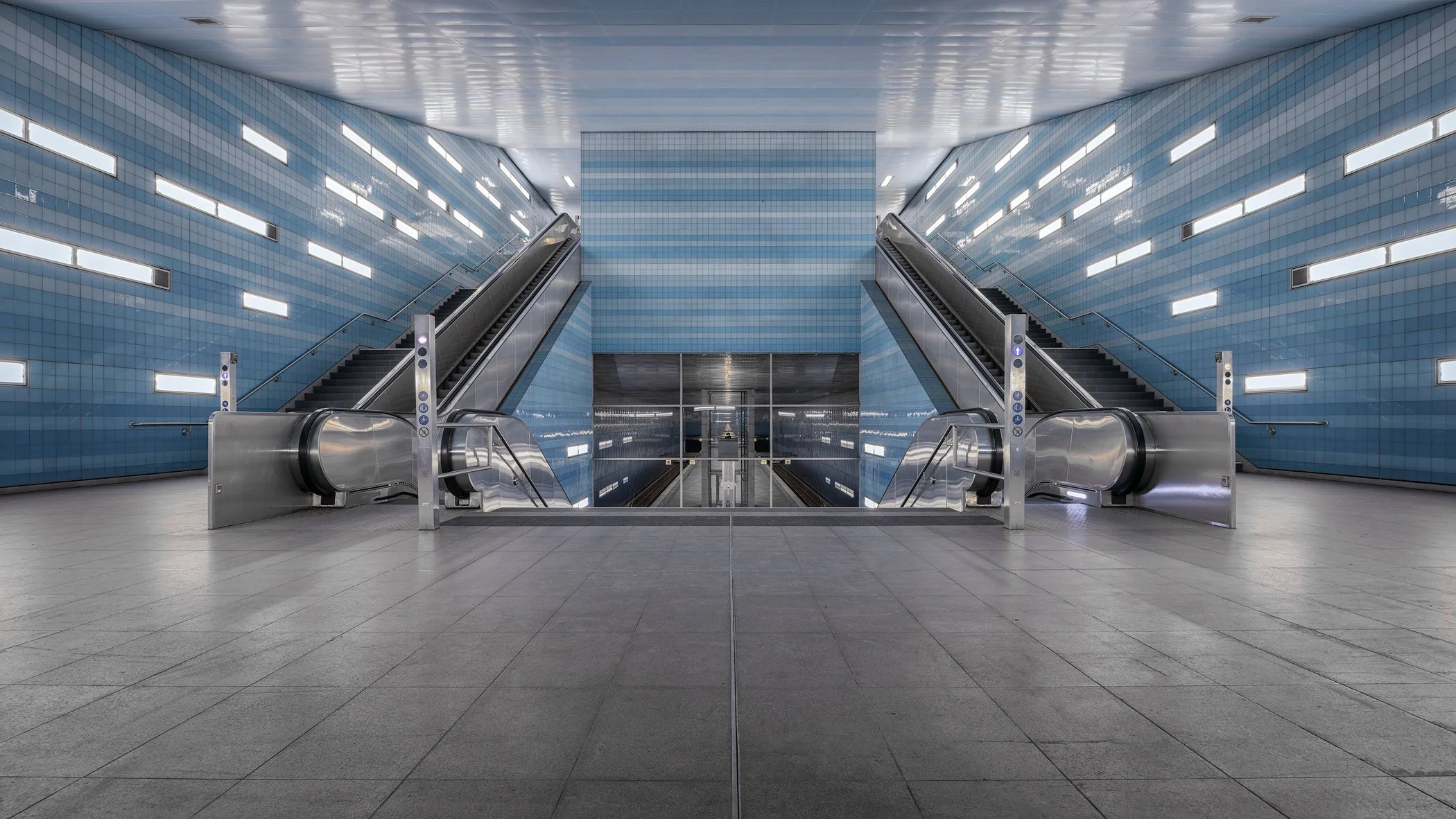 Empty underground subway station with escalators leading down, blue tiled walls, modern lighting, and reflective surfaces.