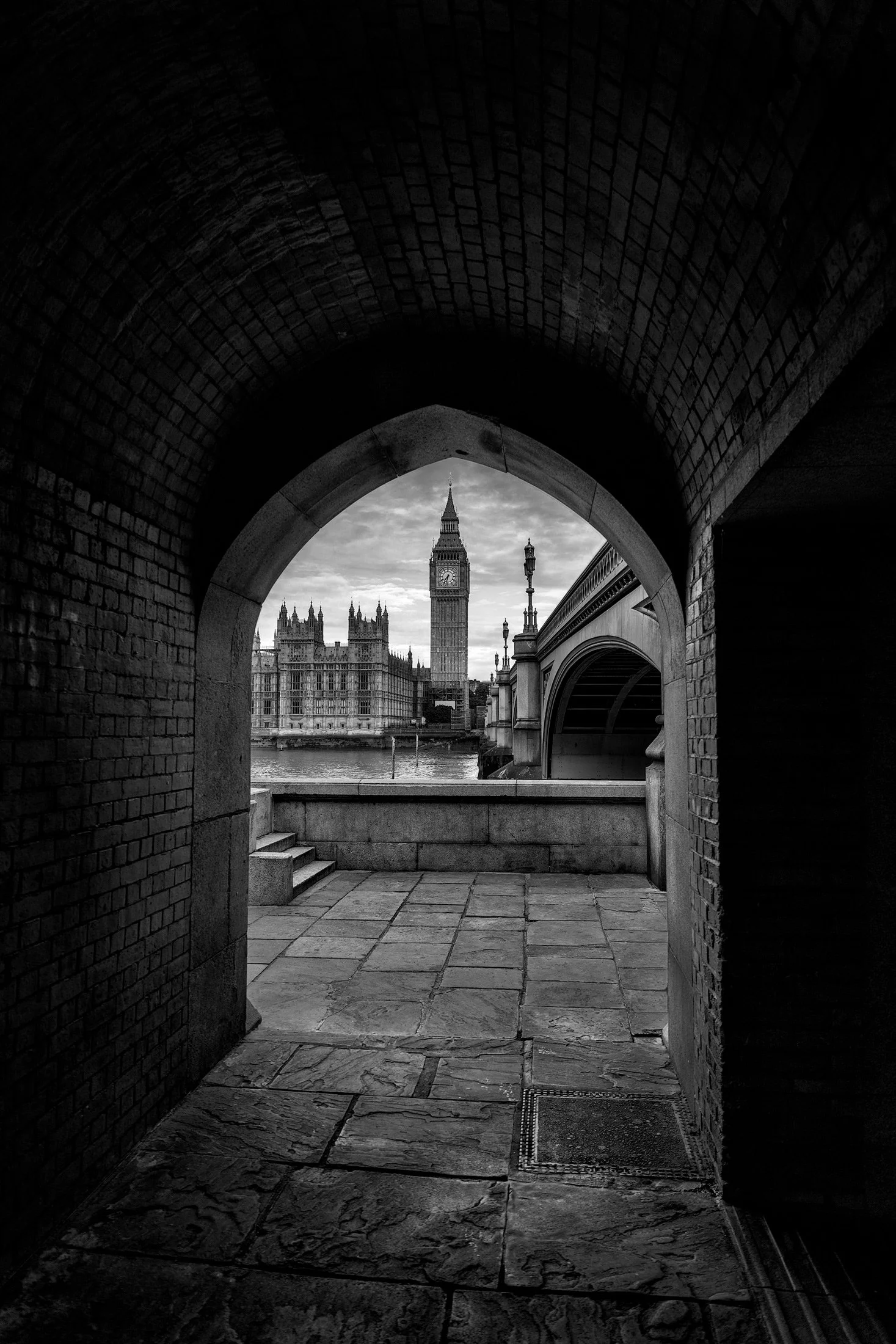 Iconic London architecture: Big Ben and the Elizabeth Tower framed through a stone archway on the South Bank. A classic monochrome view of the Palace of Westminster and Westminster Bridge over the River Thames.