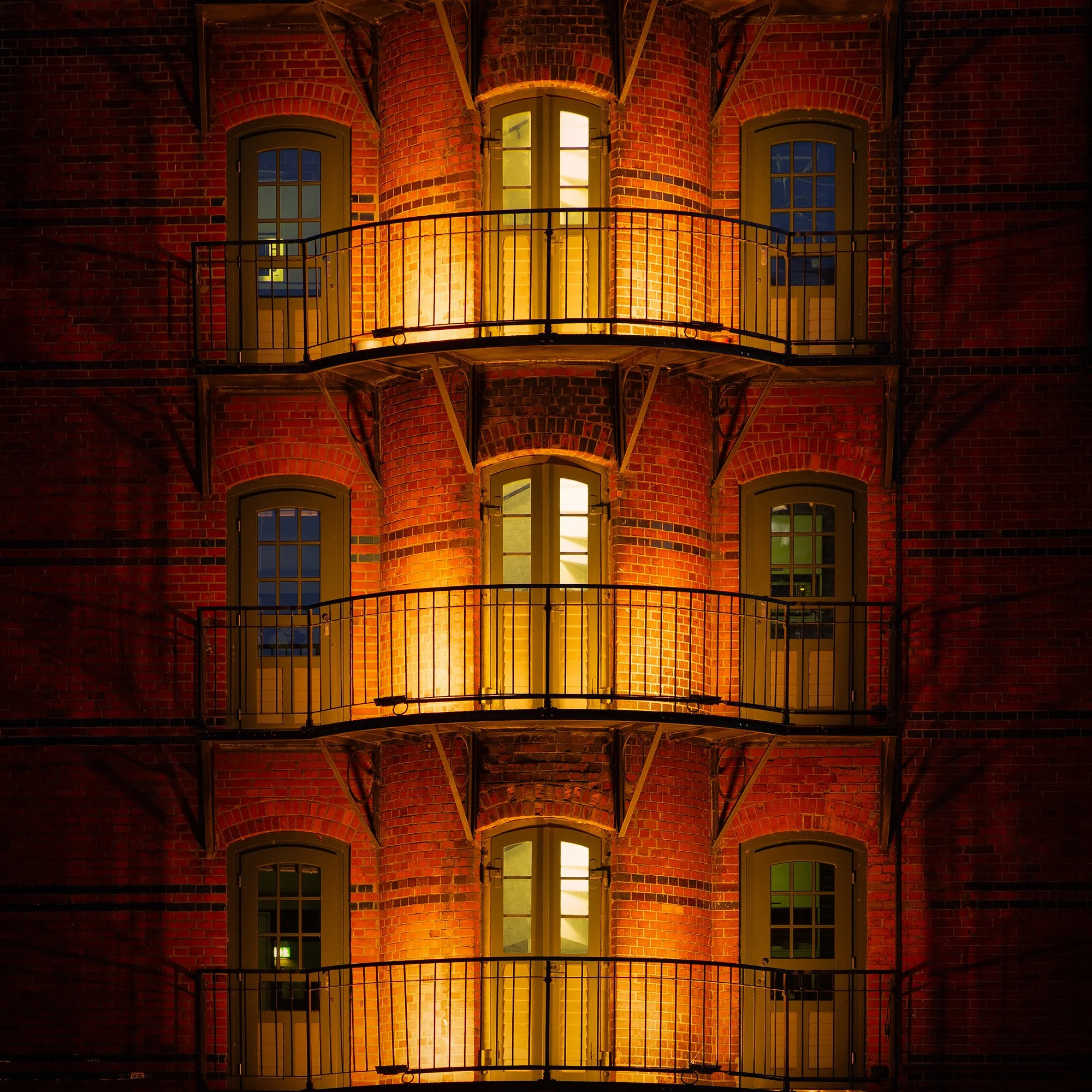Historic industrial architecture in Hamburg, Germany: A dramatic study of the Speicherstadt warehouse district. This architectural photograph captures the symmetrical, rhythmic facade of a traditional red-brick warehouse at night. The warm interior l