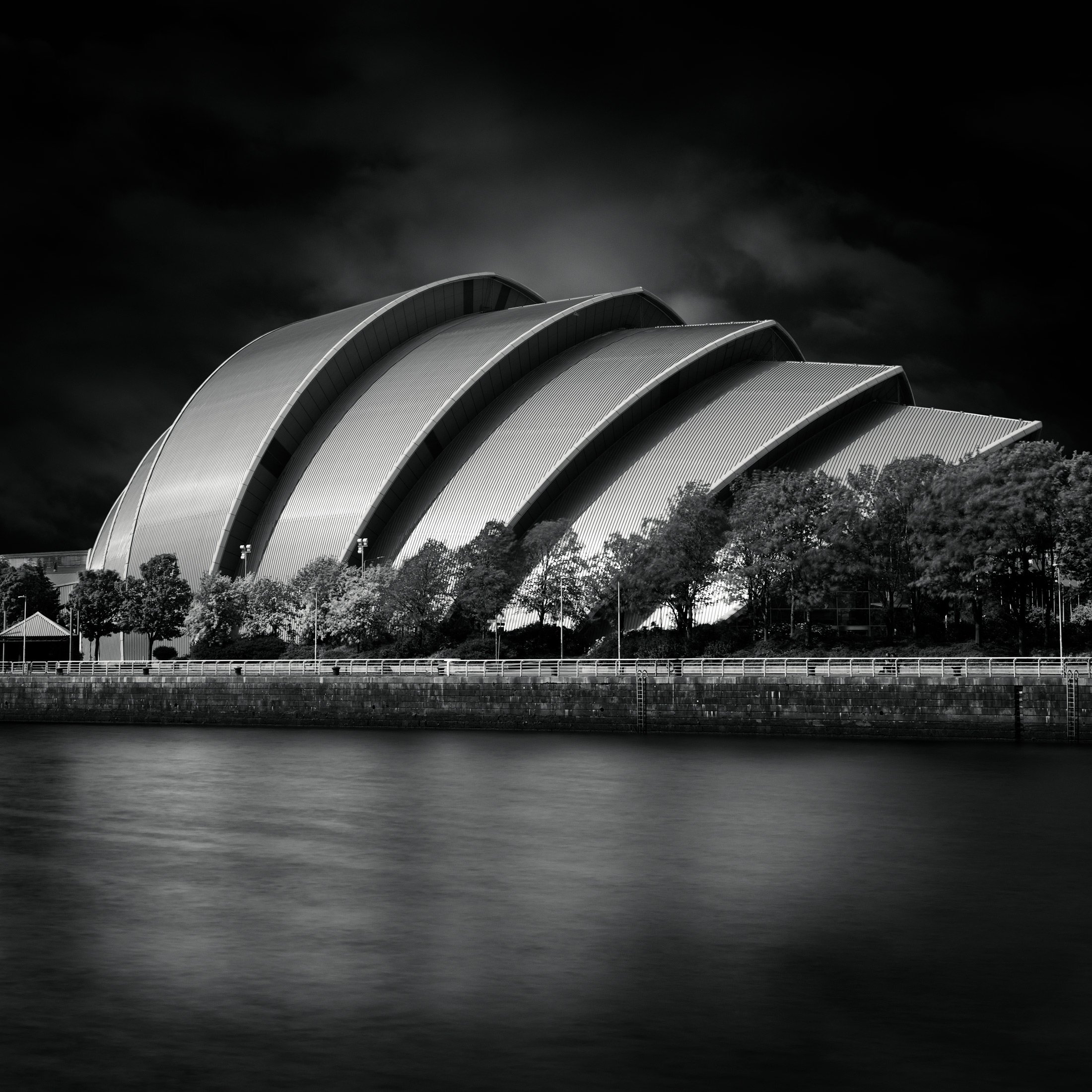 High-tech architecture in Glasgow, Scotland: The SEC Armadillo (Clyde Auditorium) designed by Foster + Partners. A cinematic black and white photograph of the iconic interlocking aluminum hulls reflected in the River Clyde under a dark, brooding sky.