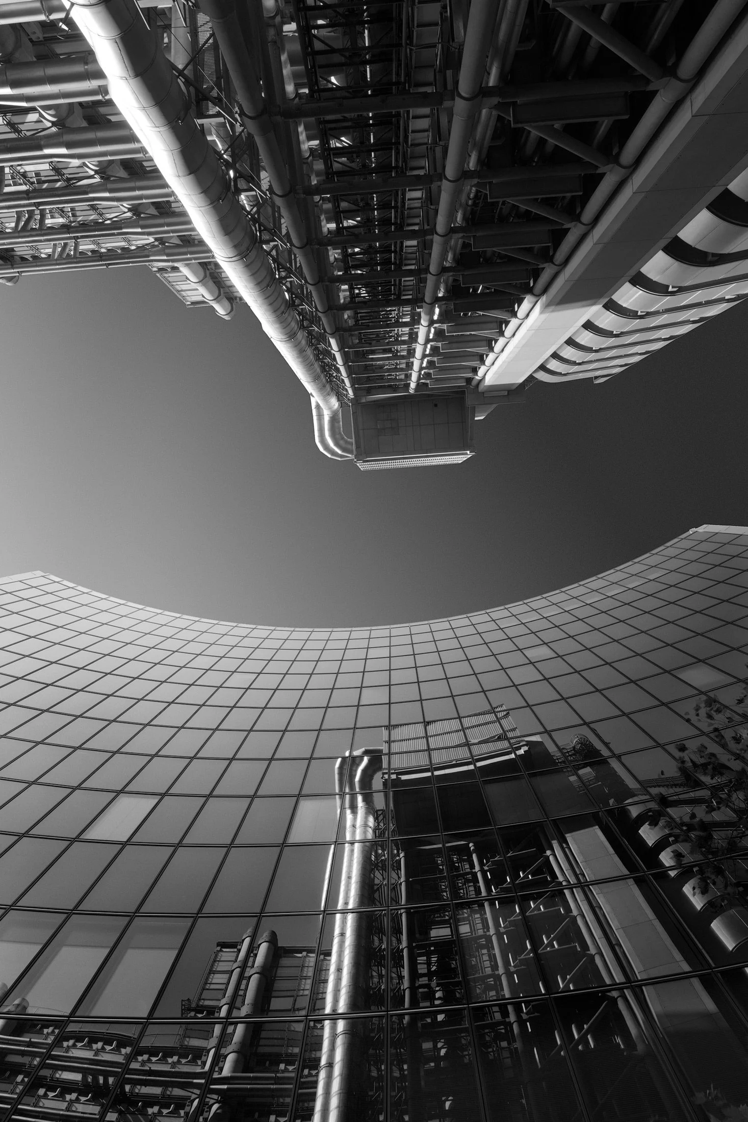 High-Tech and Modernist architecture in London: A black and white architectural study of the Lloyd’s Building reflected in the curved glass curtain wall of the Willis Building. This dramatic vertical composition highlights the contrast between Richar