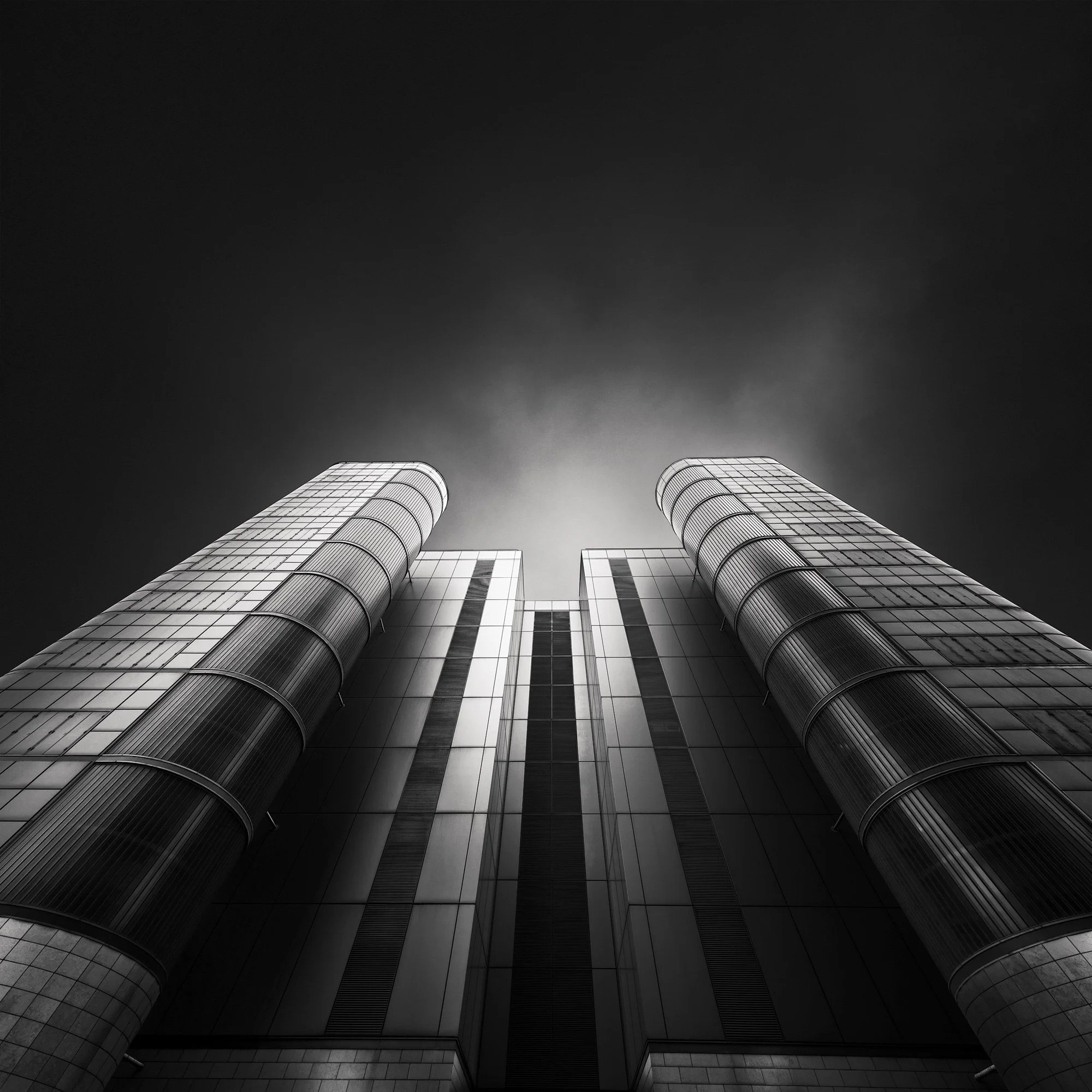 [architecture] [uk] [london] [blackandwhite] [square] Global Switch Poplar London data centre, looking up between twin rounded glass towers flanking central grid facade, dramatic dark. Architectural Photography by Jim Scott.