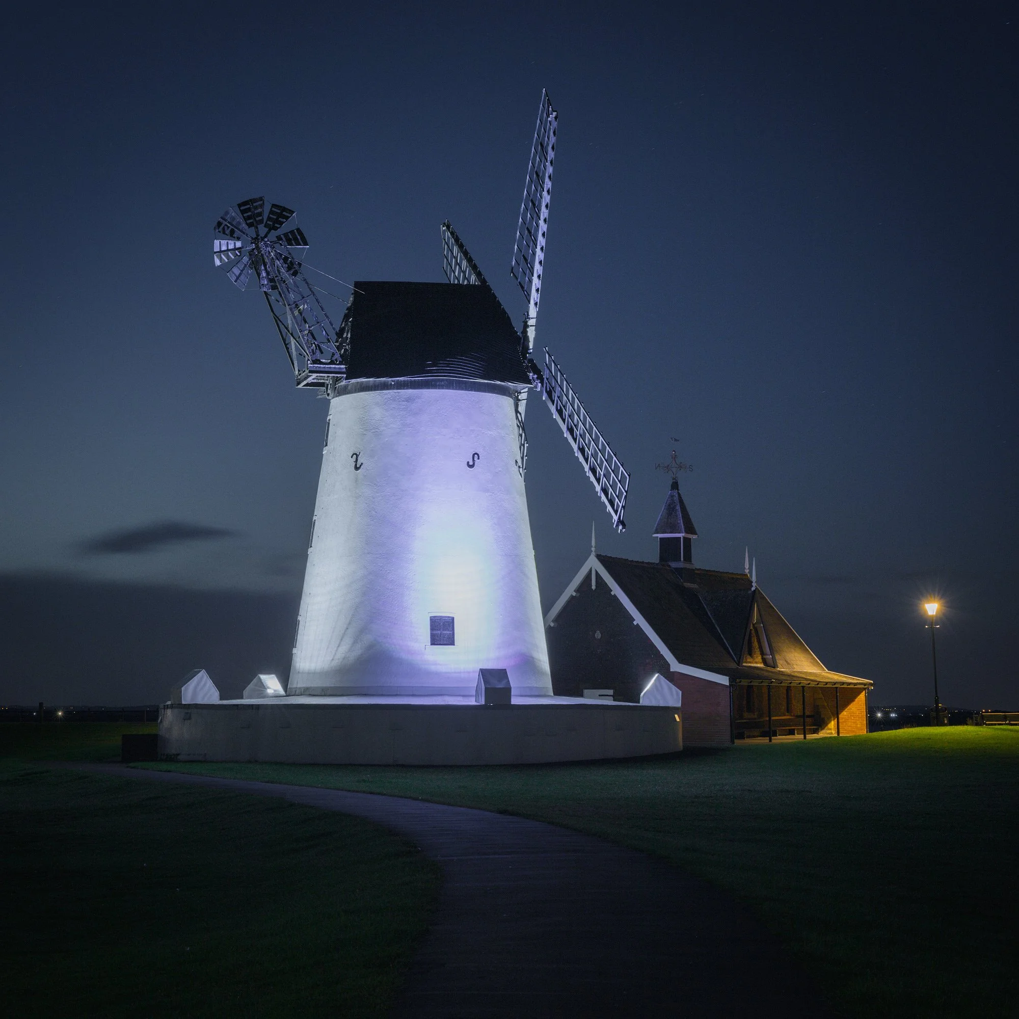 Historic industrial architecture in Lytham St Annes, UK: A dramatic study of the Lytham Windmill. This architectural photograph captures the restored 19th-century tower mill and the adjacent lifeboat house at twilight. The long-exposure technique and