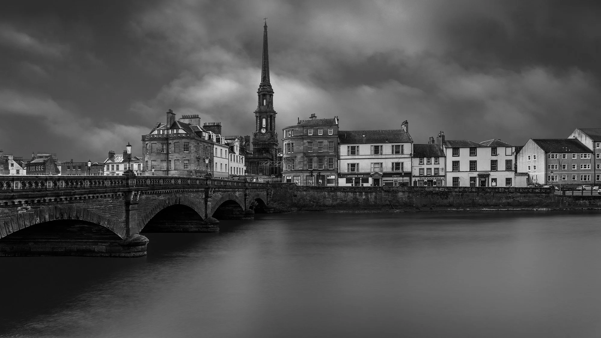 Historic Scottish urbanism in monochrome: A dramatic long-exposure study of the New Bridge of Ayr and the Town Hall spire. This architectural photograph captures the weathered sandstone textures and rhythmic arches of the bridge against a heavy, atmo