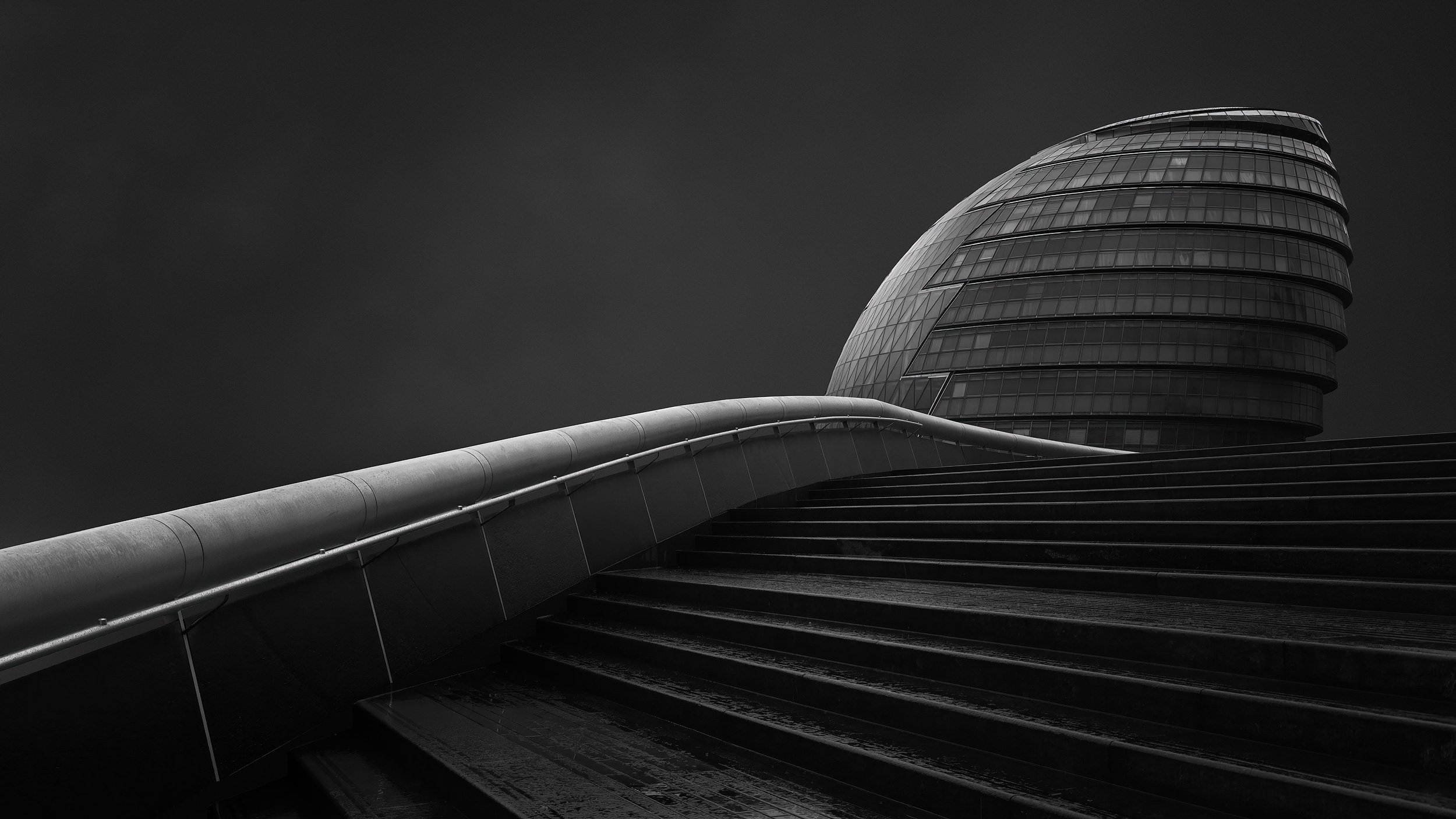 Former London City Hall from the riverside steps, tilted glass sphere leaning over a curved staircase under a dark sky. Architectural Photography by Jim Scott.