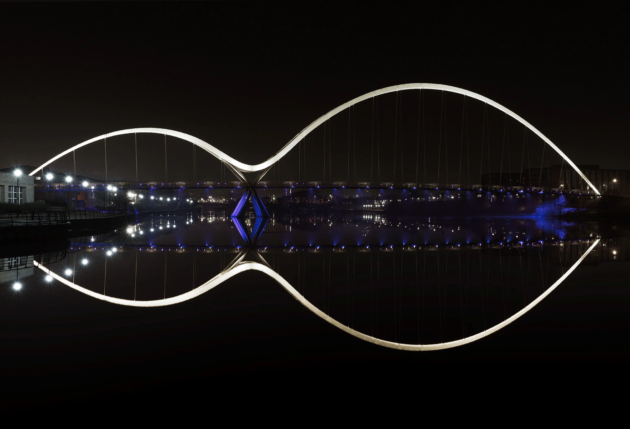 Award-winning infrastructure in Stockton-on-Tees: The Infinity Bridge at night, designed by Expedition Engineering. A symmetrical architectural photograph where the dual tied-arches and blue LED lighting reflect perfectly in the River Tees to form th