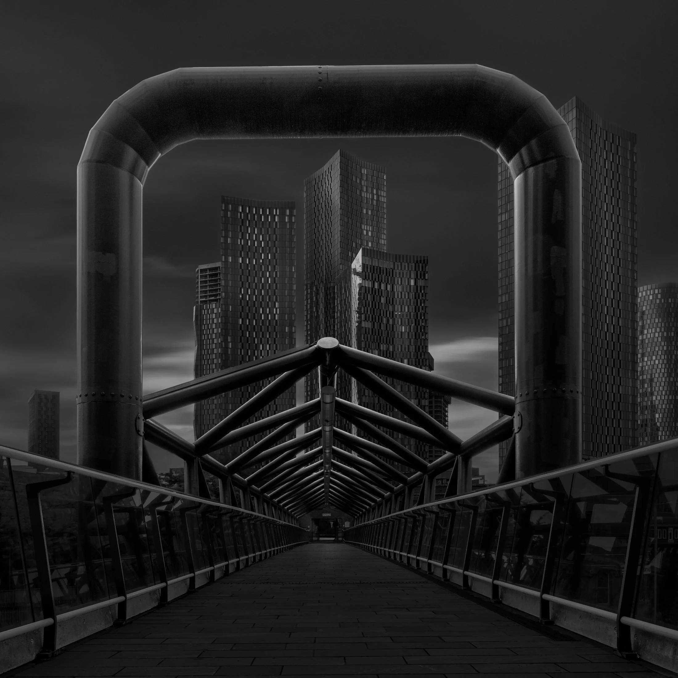 [architecture] [uk] [manchester] [blackandwhite] [square] Deansgate Station pedestrian bridge Manchester, steel pipe frame and lattice structure framing the dark tower cluster. Architectural Photography by Jim Scott.