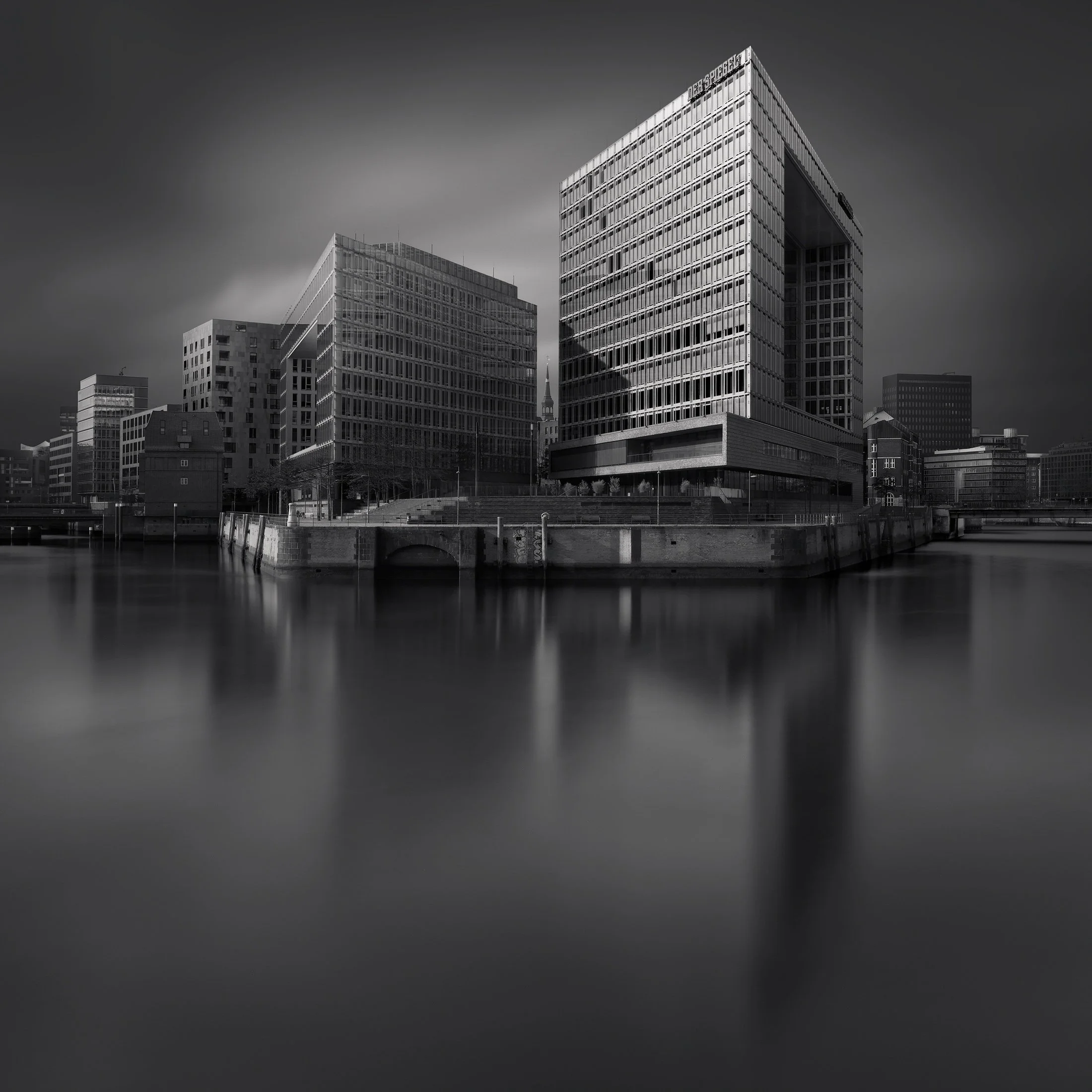 [architecture] [europe] [hamburg] [blackandwhite] [square] Der Spiegel Hamburg B&W long exposure across the canal, grid-facade block and HafenCity buildings in dark water. Architectural Photography by Jim Scott.