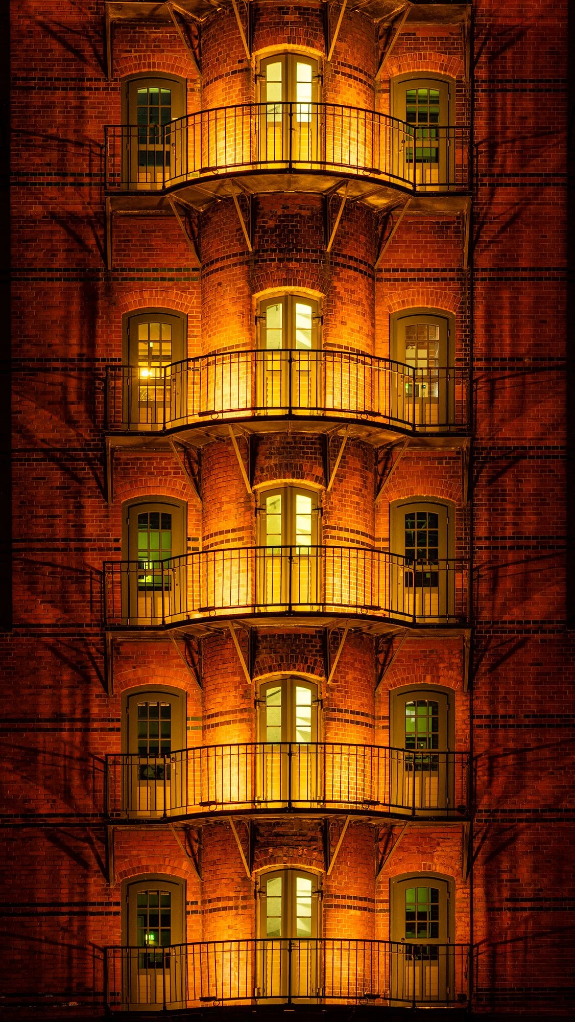 Gothic Revival architecture in Hamburg, Germany: A symmetrical night study of a red-brick warehouse facade in the historic Speicherstadt district. This vibrant colour photograph highlights the warm glow of illuminated arched windows and repeating cur