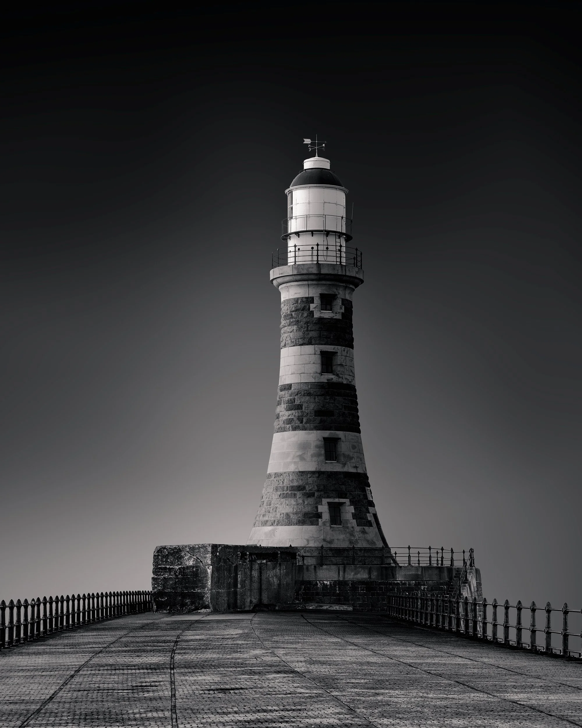 Historic maritime architecture in Sunderland, UK: A dramatic monochromatic study of the Roker Pier Lighthouse. This vertical architectural photograph captures the lighthouse’s distinctive banded granite tower and lantern house at the end of the long 