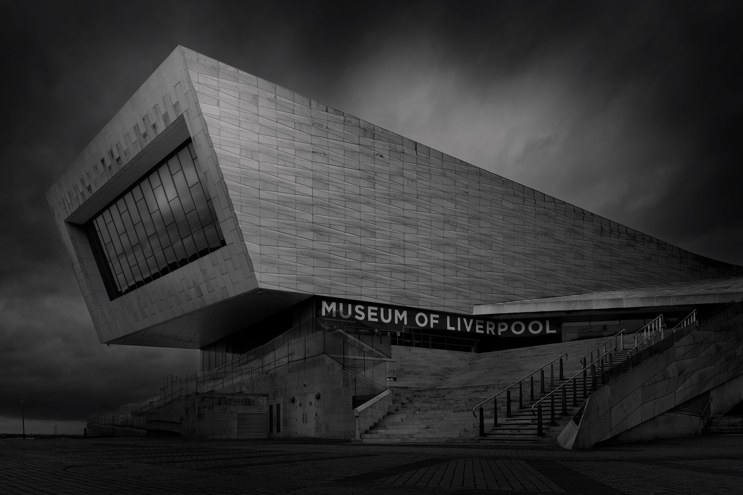 Deconstructivist architecture in Liverpool, UK: A dramatic monochromatic study of the Museum of Liverpool, designed by 3XN. This architectural photograph captures the building’s aggressive, cantilevered geometry and textured limestone facade against 