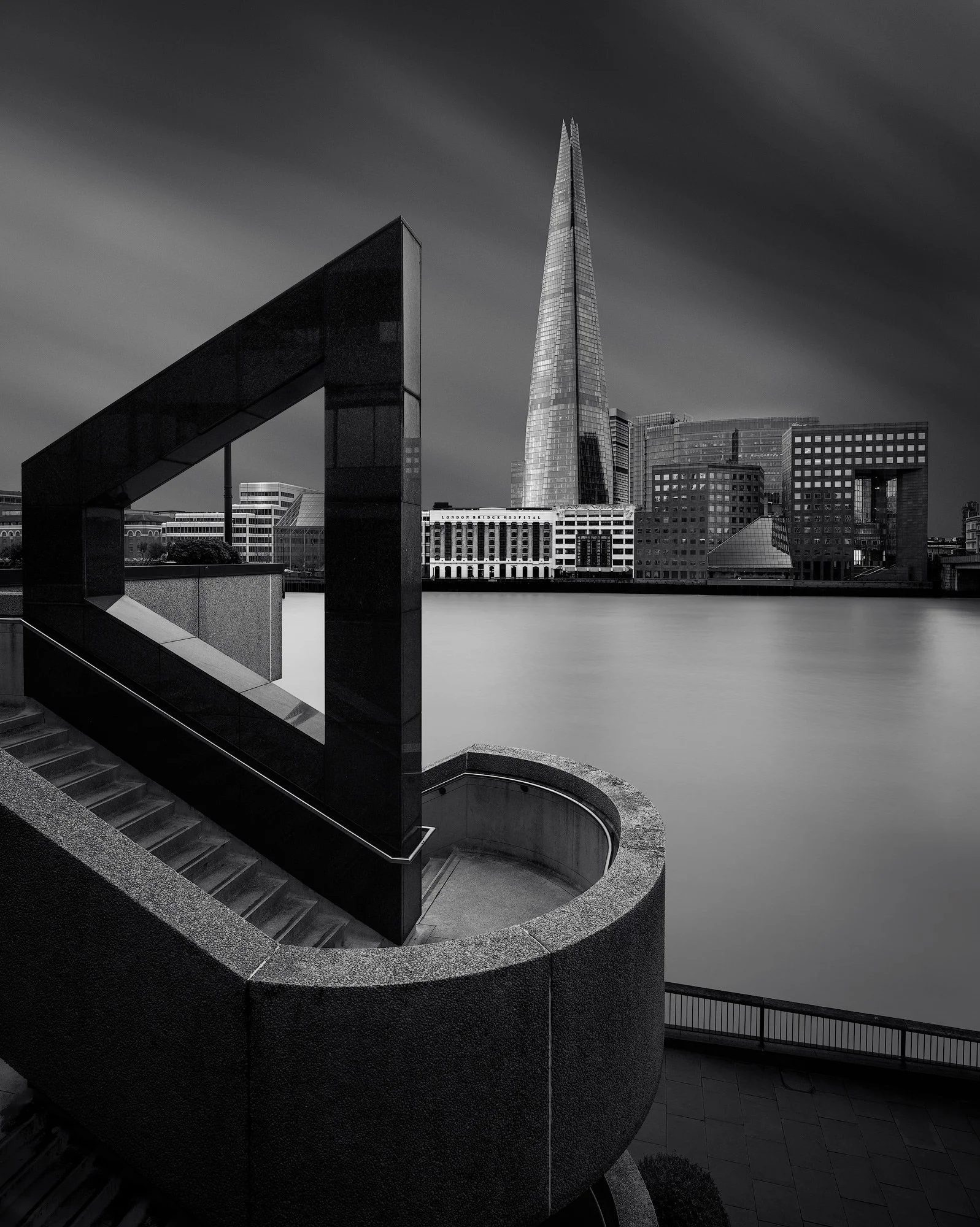 Contemporary urban geometry in London Bridge: A framed monochromatic study of The Shard, designed by Renzo Piano. This architectural photograph utilises the sharp, geometric granite structures of the South Bank to create a dramatic foreground frame f