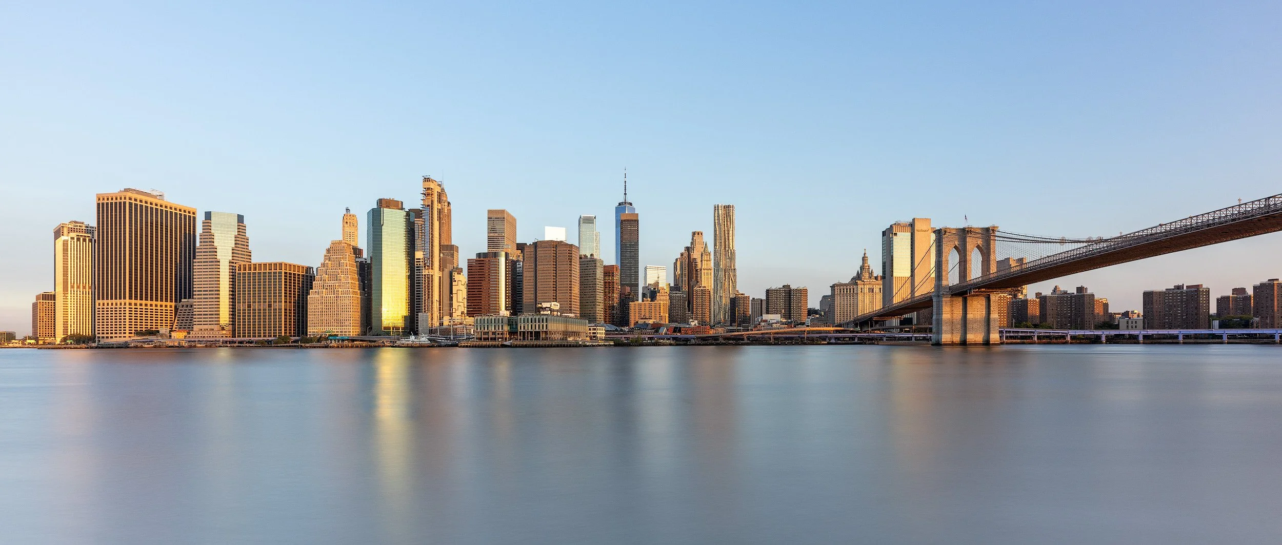 Contemporary and historic commercial architecture in Manhattan, New York: A dramatic study of the Lower Manhattan skyline and the Brooklyn Bridge at golden hour. This panoramic architectural photograph captures the dense cluster of financial district