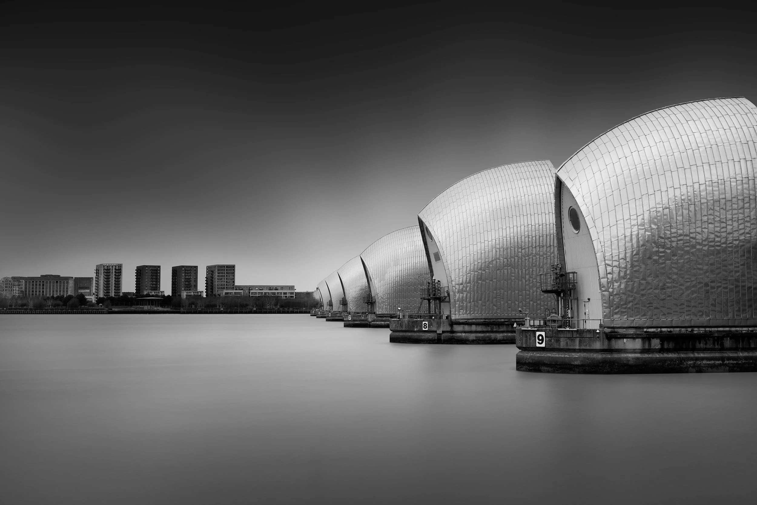 Iconic civil engineering in London, UK: A dramatic monochromatic study of the Thames Barrier, designed by Rendel, Palmer and Tritton. This long-exposure photograph captures the rhythmic recession of the stainless-steel flood gates across the river. T