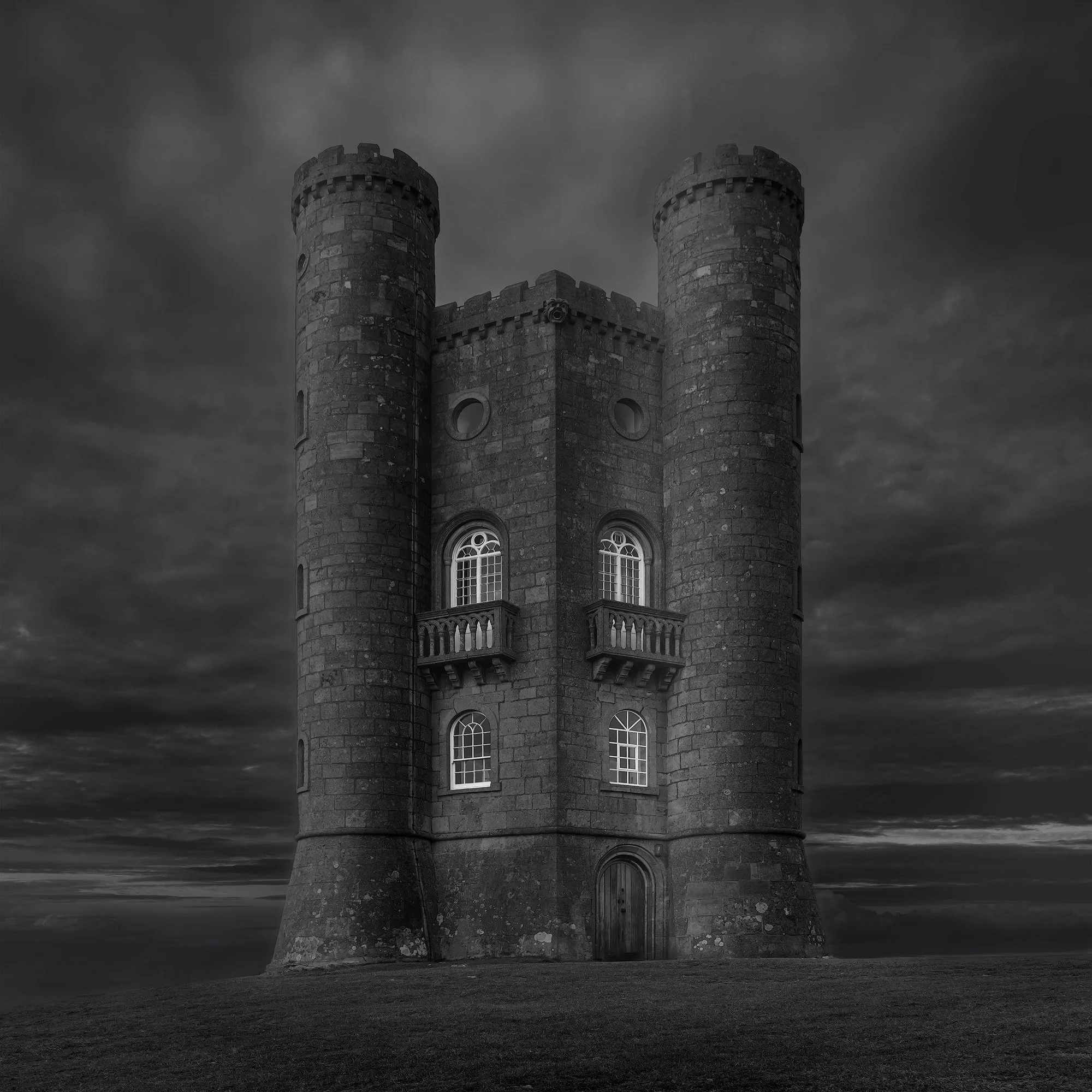18th-century Gothic Folly in the Cotswolds, UK: A dramatic monochromatic study of Broadway Tower in Worcestershire. This architectural photograph captures the tower's symmetrical turrets and weathered stone textures against a brooding, atmospheric sk