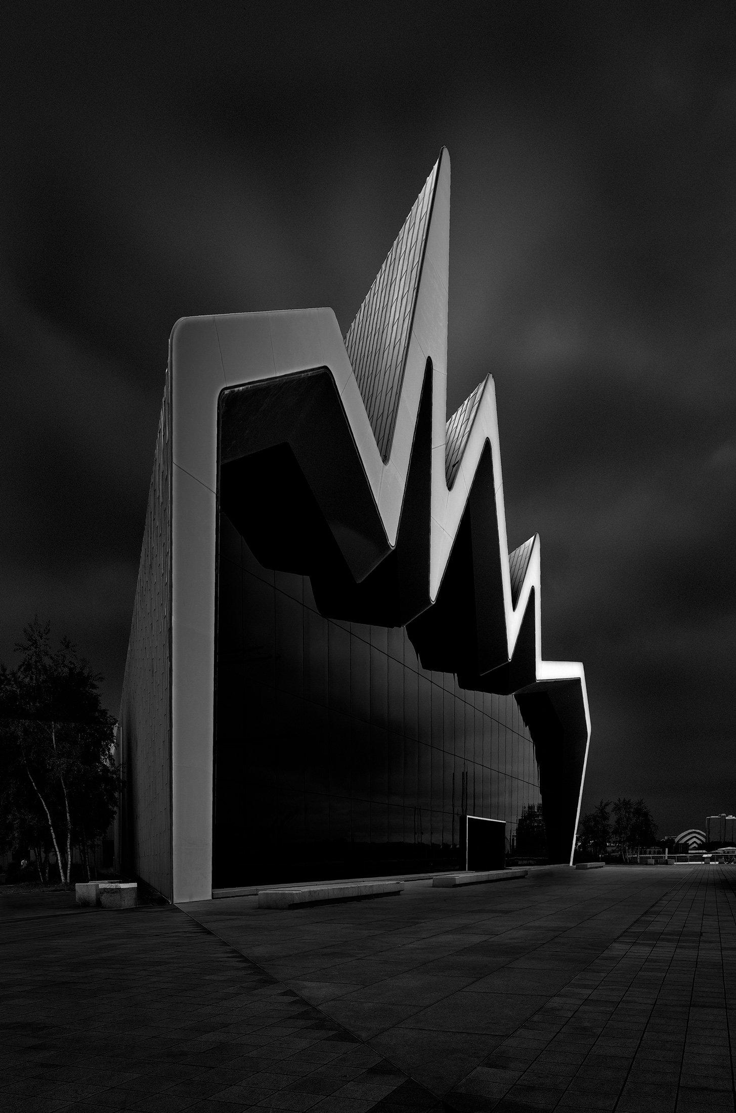 Contemporary architecture in Glasgow, Scotland: A dramatic monochromatic study of the Riverside Museum, designed by Zaha Hadid. This architectural photograph captures the building’s aggressive, zigzag roofline and textured zinc cladding against a moo