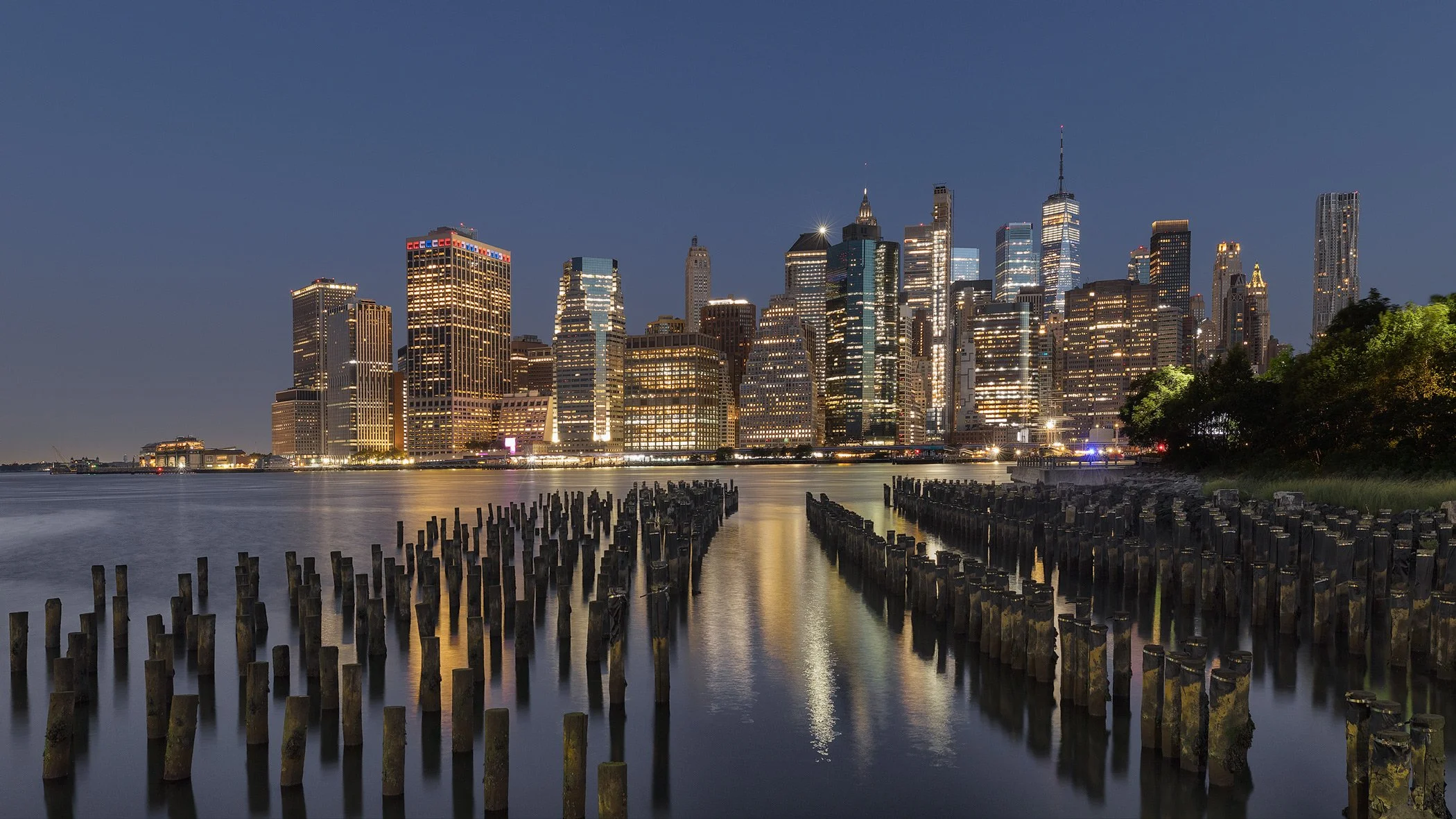 [architecture] [global] [newyork] [colour] [panoramic] [nightscape] Blue hour panorama of Lower Manhattan from Brooklyn Bridge Park, timber pier pilings leading to glittering towers. Architectural Photography by Jim Scott.