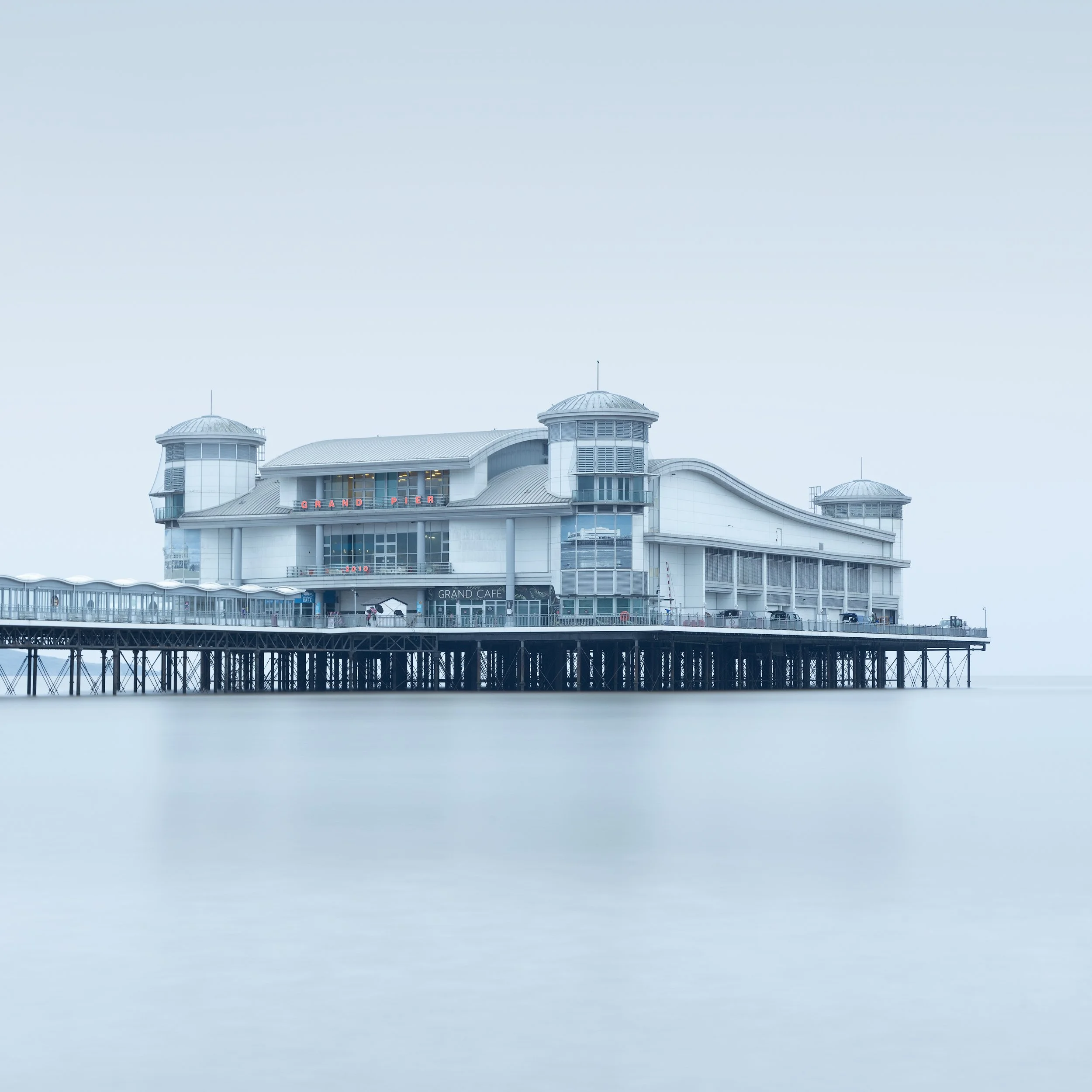 Contemporary coastal architecture in Somerset, UK: A high-key minimalist study of the Grand Pier at Weston-super-Mare, redesigned by Angus Meek Architects. This long-exposure photograph captures the pavilion’s symmetrical towers and curved roofline f