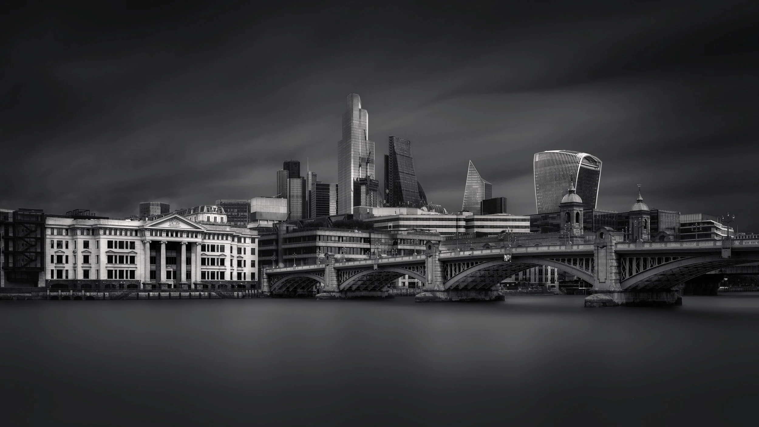 [architecture] [uk] [london] [blackandwhite] [panoramic] London skyline B&W long exposure from Bankside, Southwark Bridge and City towers rising above a silken smooth Thames. Architectural Photography by Jim Scott.