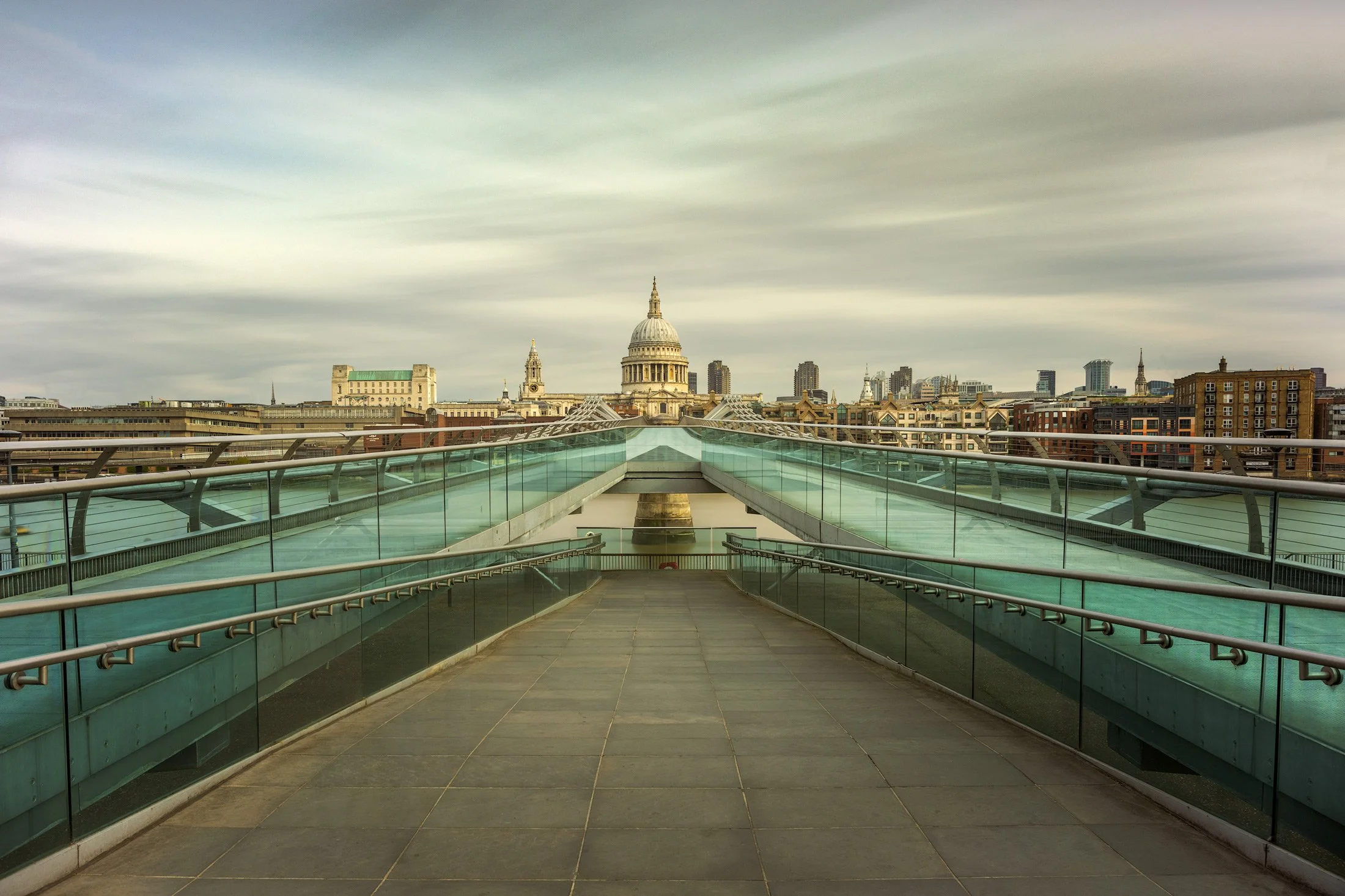 Iconic London skyline at Bankside: A central perspective of the Millennium Bridge looking towards St Paul’s Cathedral. This colour architectural photograph uses a long-exposure to create a soft, ethereal sky and water, highlighting the teal-tinted gl