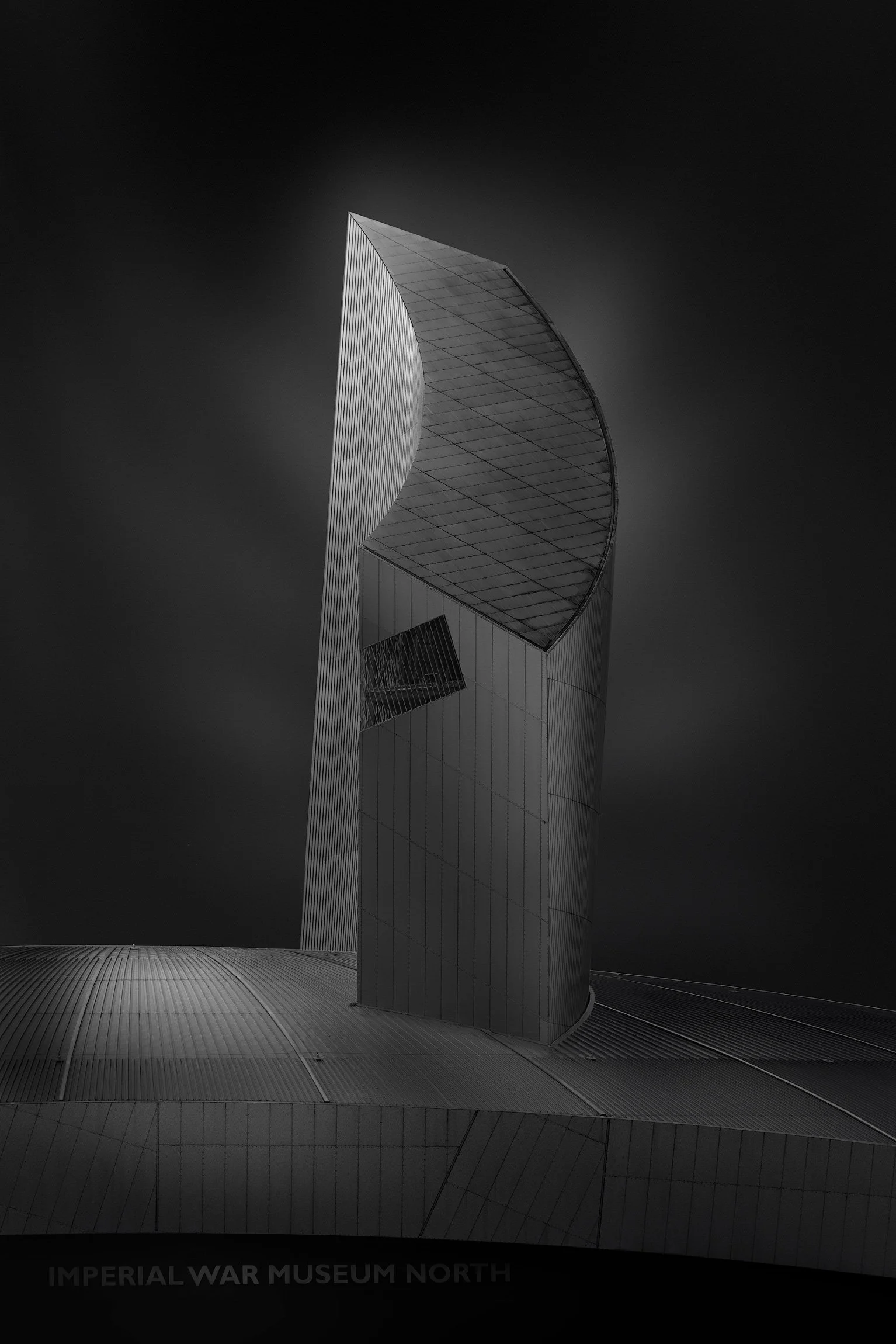 Architecture in Salford, UK: A dramatic monochromatic study of Imperial War Museum North, designed by Daniel Libeskind. This architectural photograph captures the "Air Shard," a soaring, vertical aluminium-clad structure that curves against a dark, a