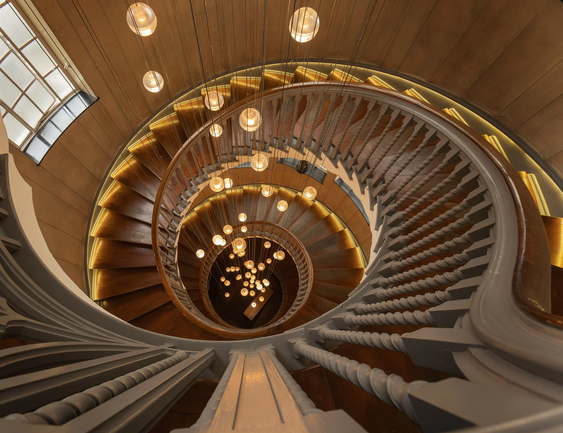 Historic Edwardian interior design in London, UK: A symmetrical downward study of the iconic Brewer Staircase at Heal’s. This photograph captures the sweeping, mathematical spiral of the wooden steps and the rhythmic white balustrades. The warm, radi