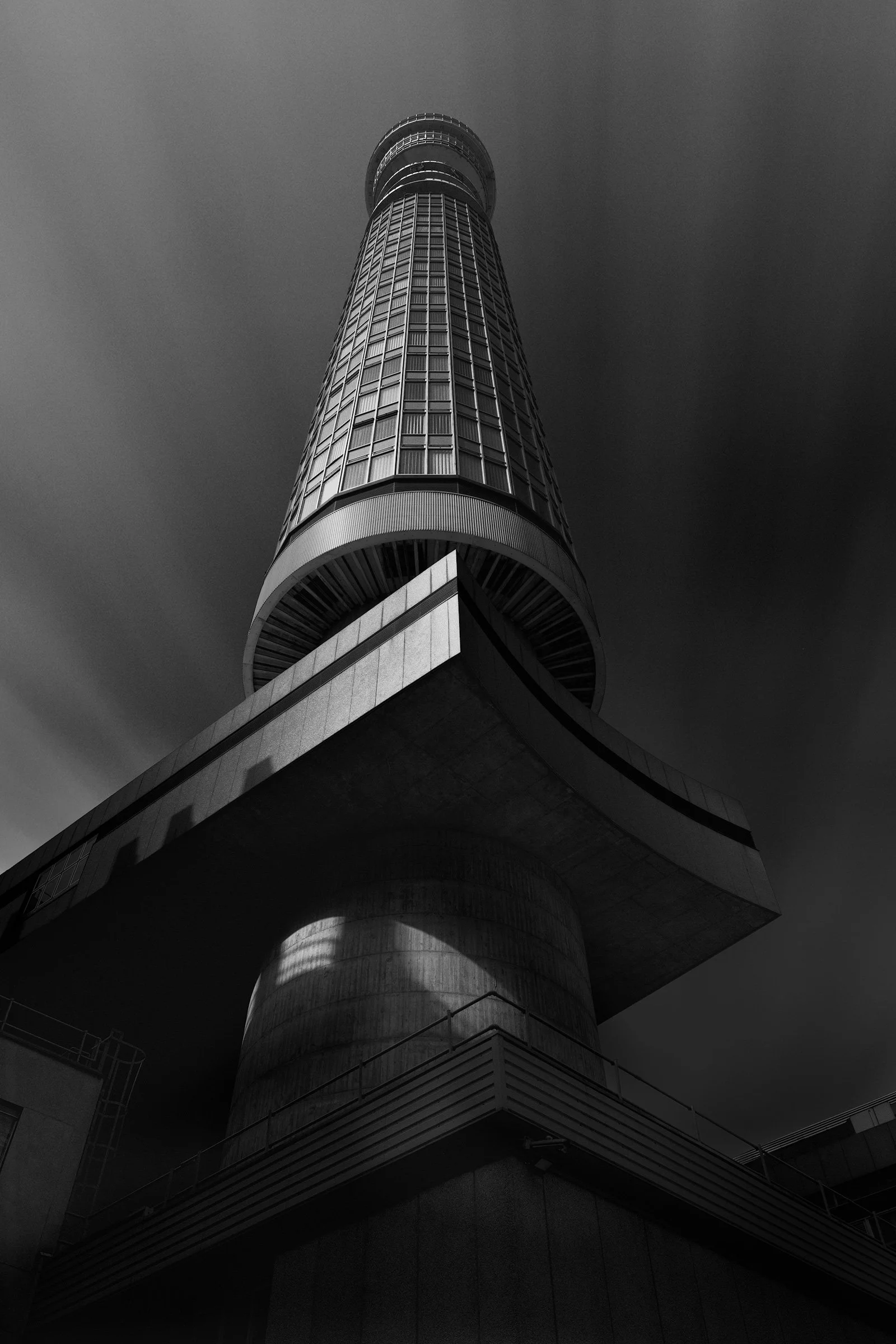 Mid-century modern infrastructure in London, UK: A dramatic monochromatic study of the BT Tower, viewed from a low-angle perspective. This architectural photograph captures the building's iconic cylindrical form and brutalist concrete base against a 