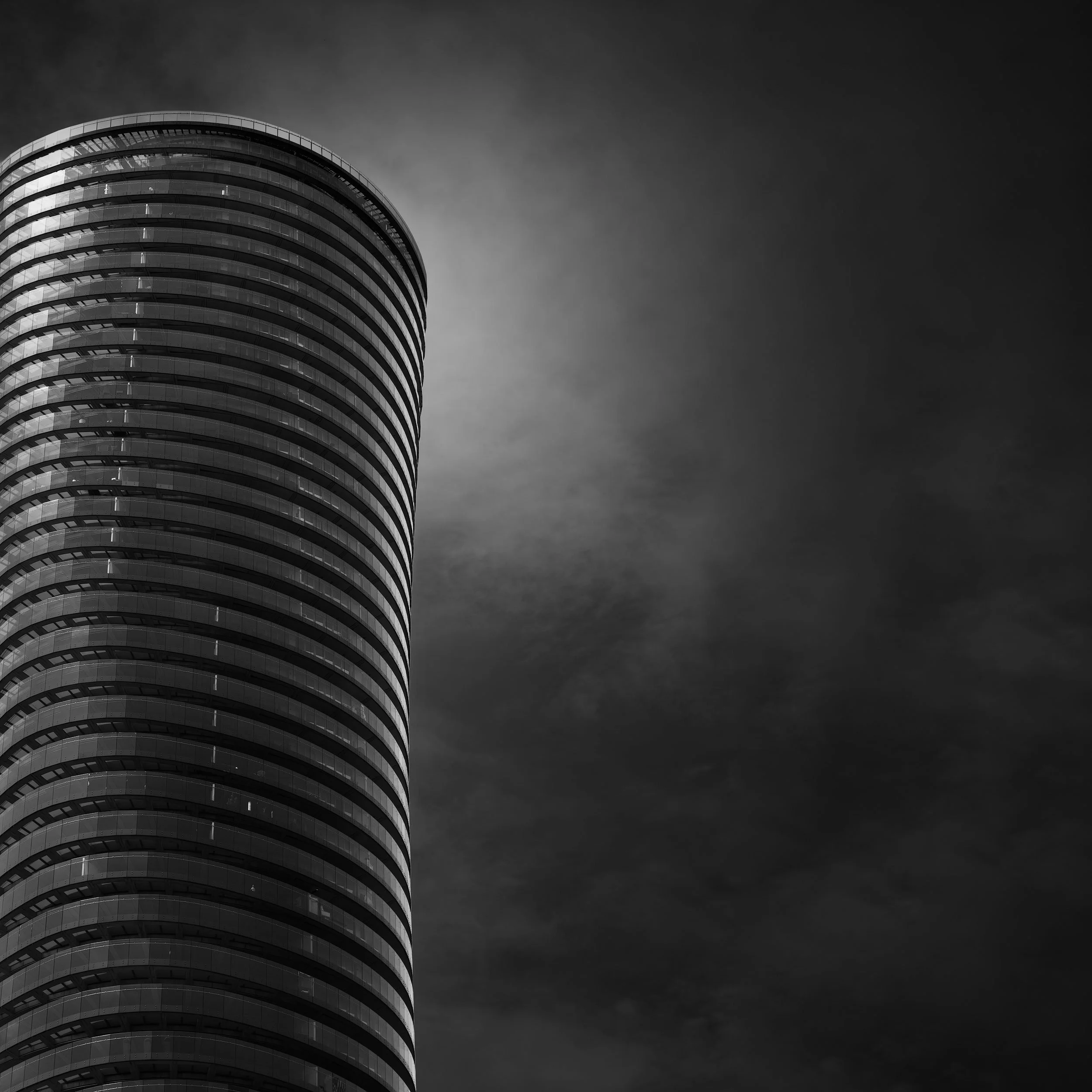 [architecture] [uk] [london] [blackandwhite] [square] Arena Tower London monochrome, cylindrical horizontally banded form against a brooding dark storm-filled sky. Architectural Photography by Jim Scott.