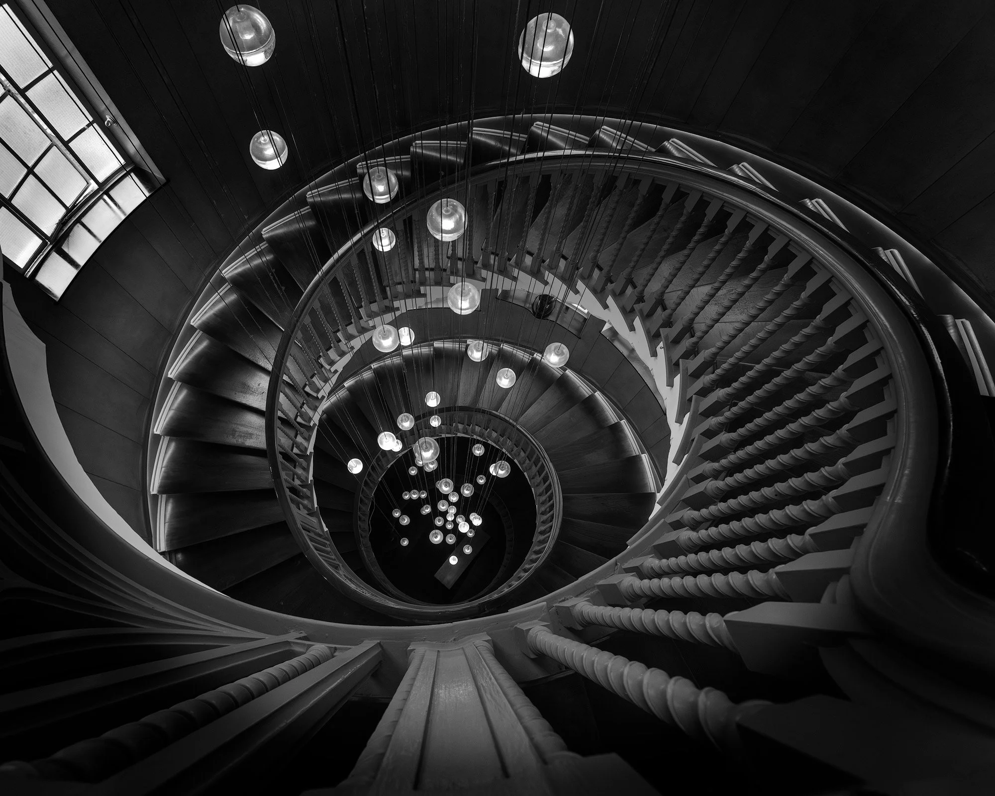 Edwardian architectural craftsmanship in London, UK: A symmetrical monochromatic top-down study of the Cecil Brewer Staircase at Heal’s. This interior photograph captures the perfect geometric spiral of the wooden banister and the rhythmic repetition