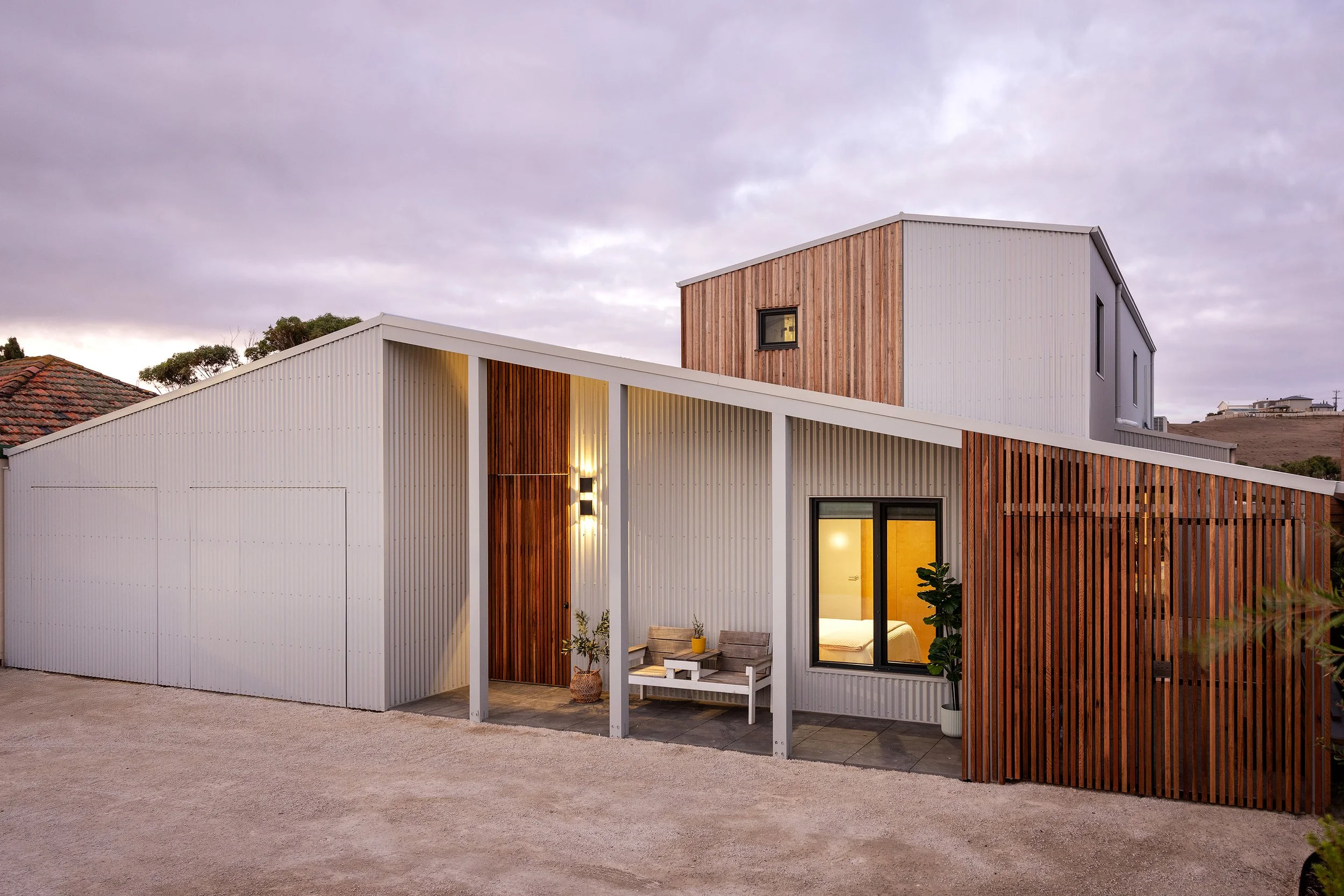 Modern coastal architecture in Port Fairy at twilight, showing a white and timber exterior home with large windows connecting to an outdoor seating area. Designed for the Great Ocean Road climate. PassivHaus
