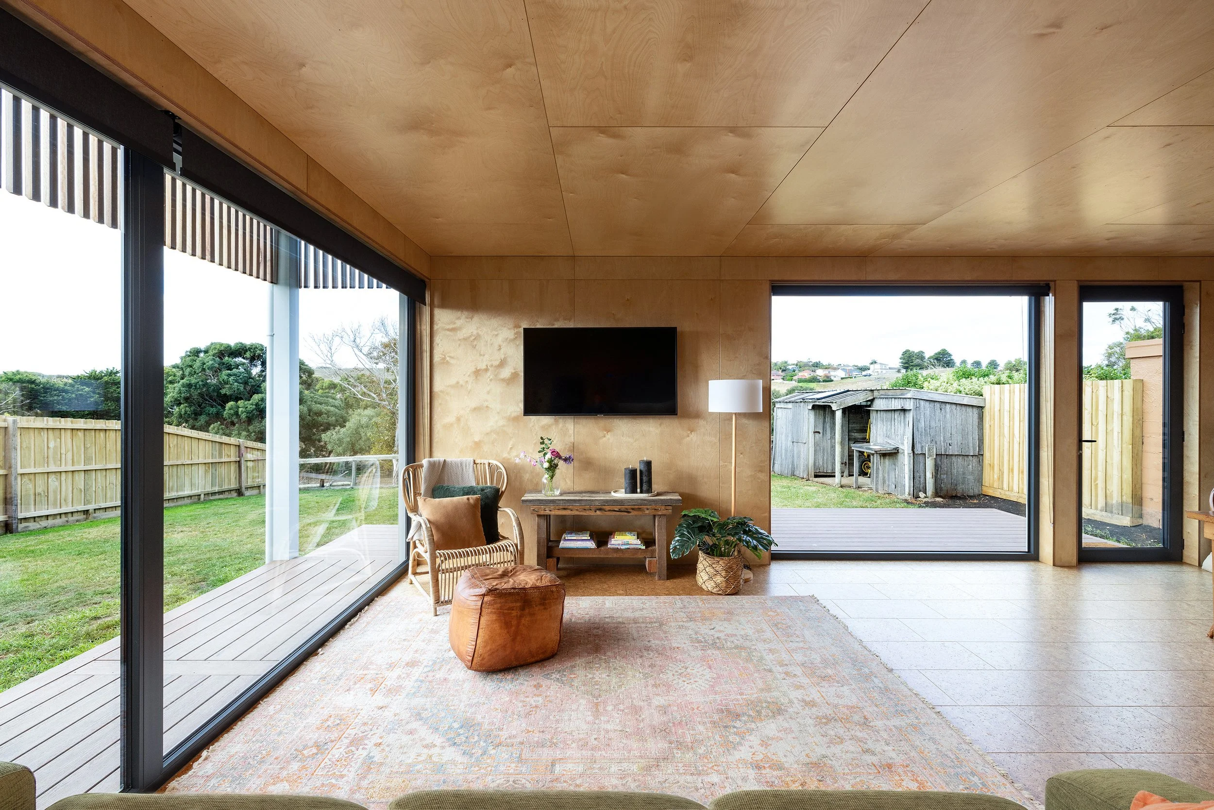 Living room with large sliding glass doors, wooden ceiling, beige walls, flat-screen TV mounted on the wall, wicker chair with cushions, wooden table with flowers and candles, potted plant, area rug, and a view of a backyard with a wooden shed and garden.
