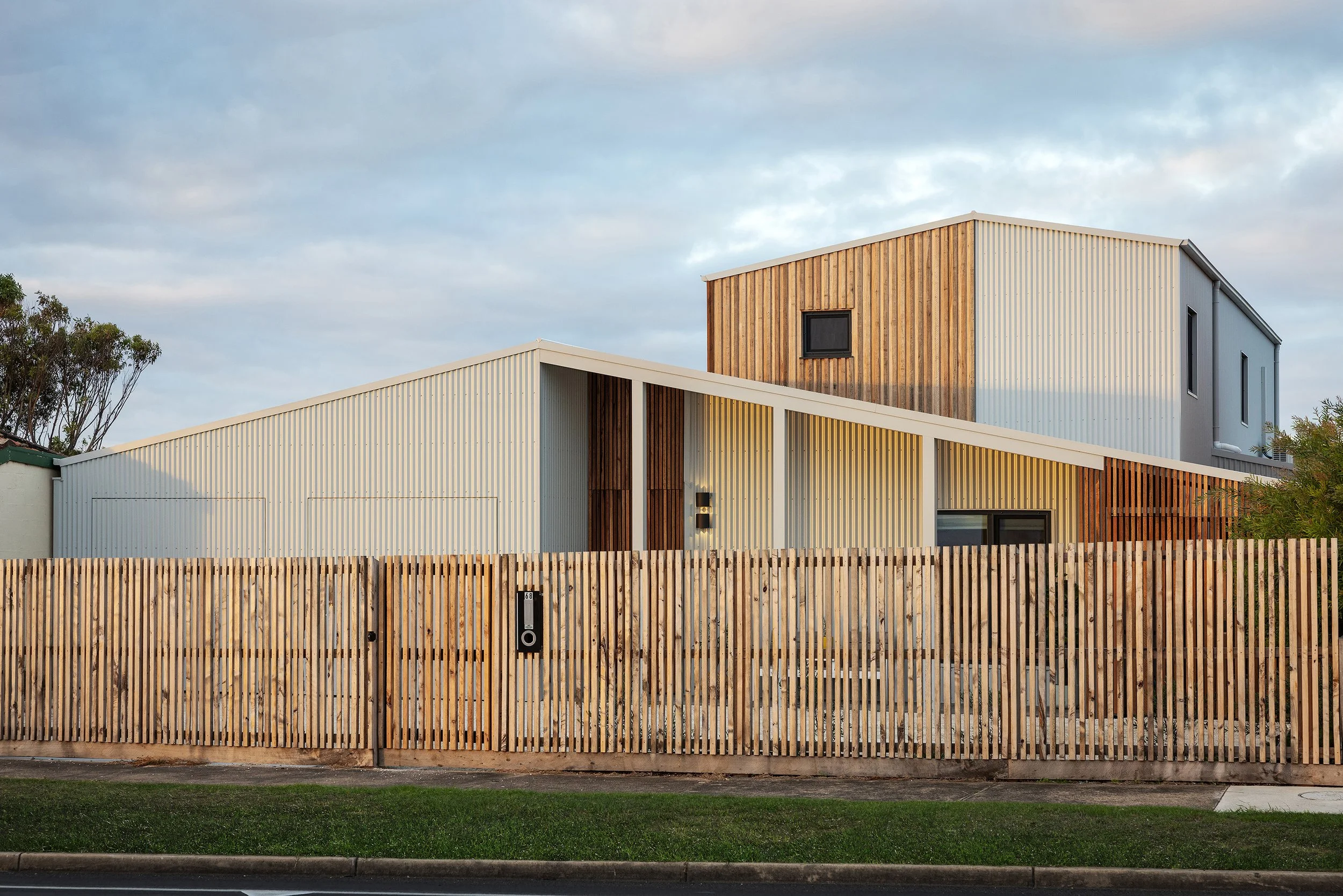 Modern passivhaus with a combination of wood and metal exterior panels, surrounded by a wooden fence and a grassy lawn, under a cloudy sky.