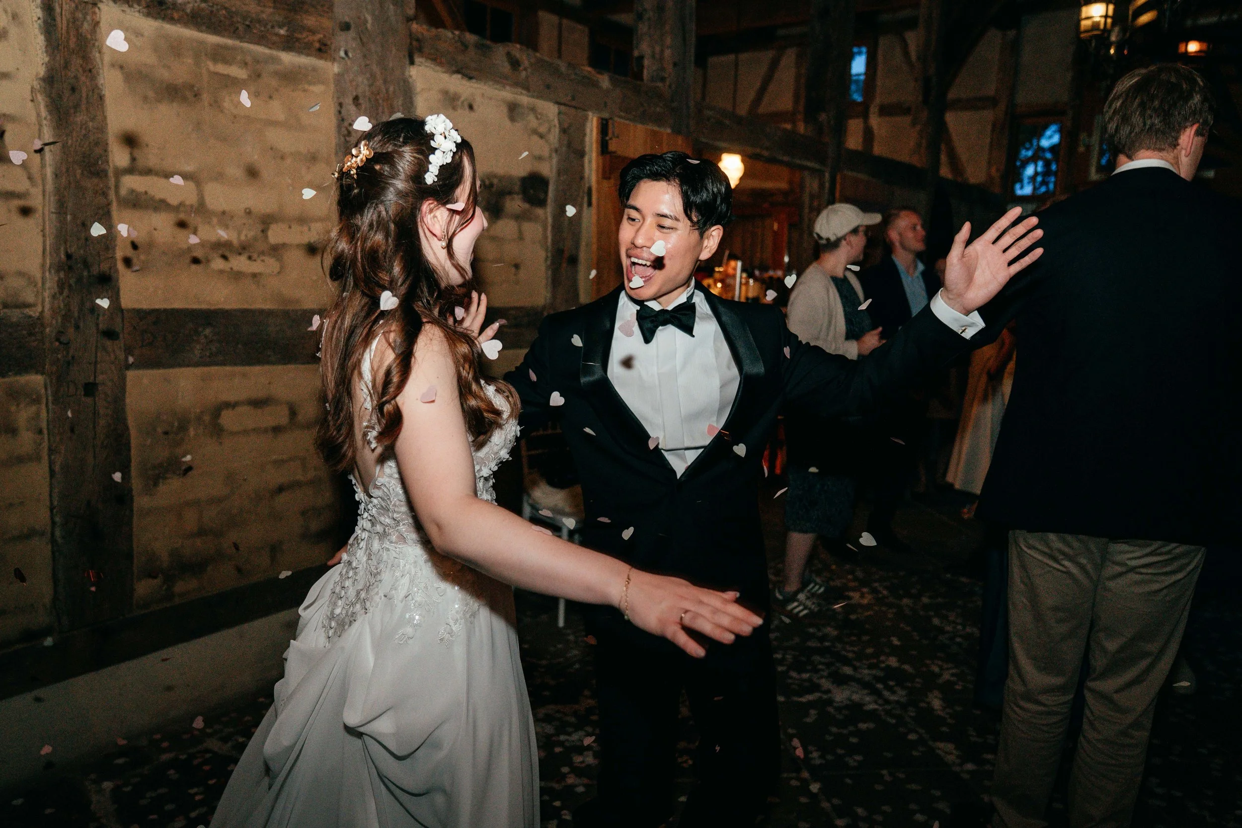 Ein älteres Paar tanzt bei einer Hochzeit in einem rustikalen Raum, umgeben von Freunden und Konfetti.