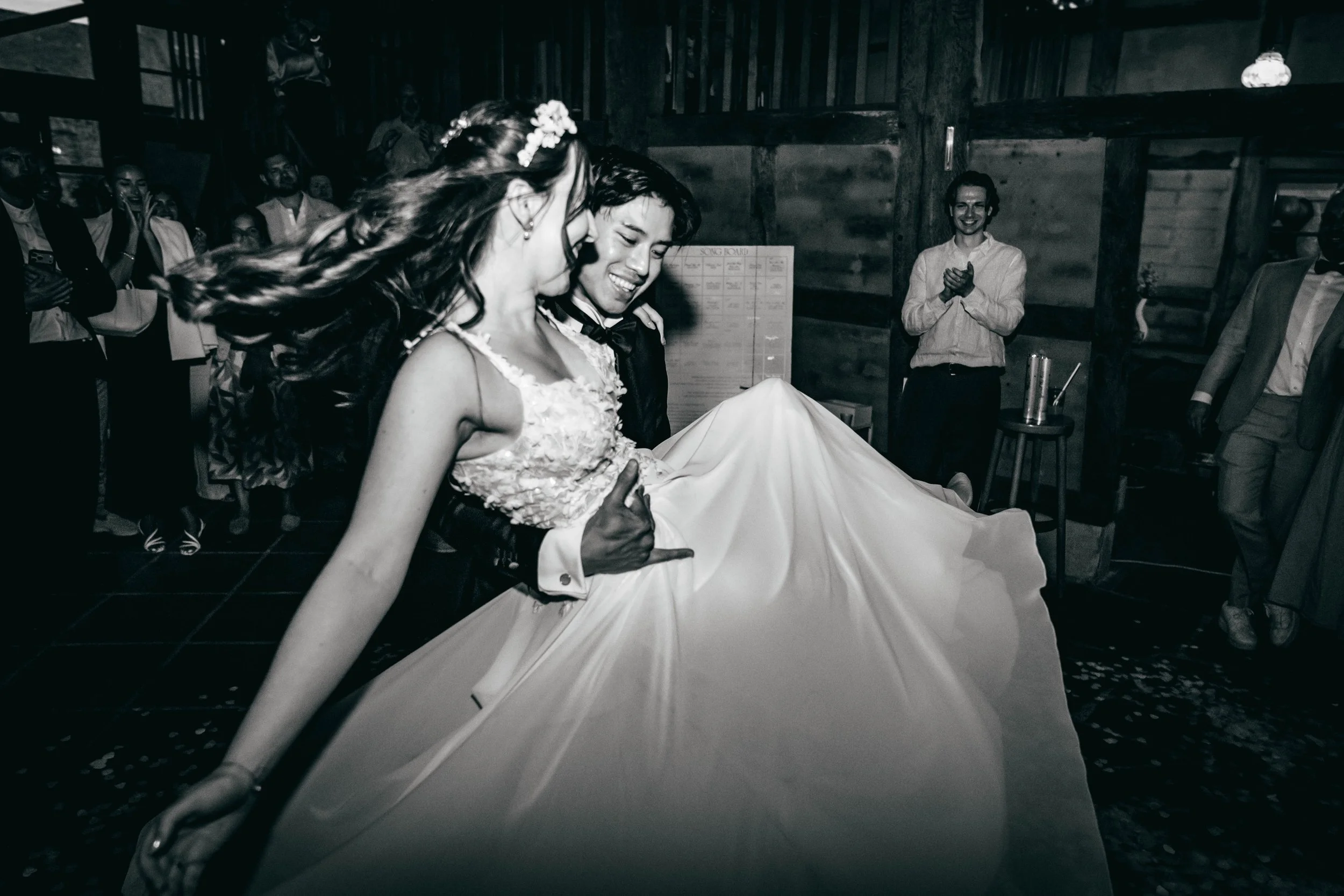 Ein Brautpaar tanzt bei einer Hochzeit, umgeben von lachenden Gästen in einem rustikalen Holzgebäude.