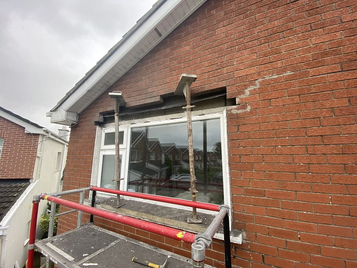 Brick house exterior with window undergoing repairs, scaffolding in front, and cloudy sky.
