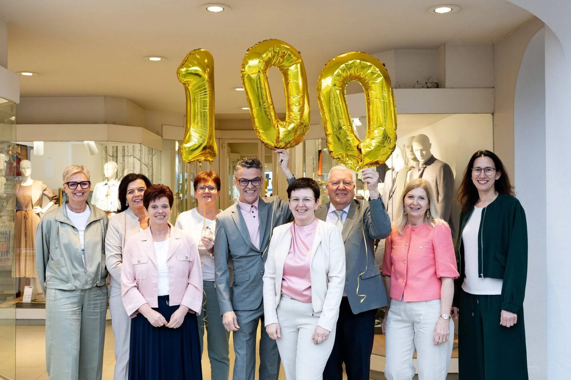 Die Belegschaft des Modehaus FRANK  stehen vor dem Geschäft und halten Luftballons in Form der Zahl 100.
