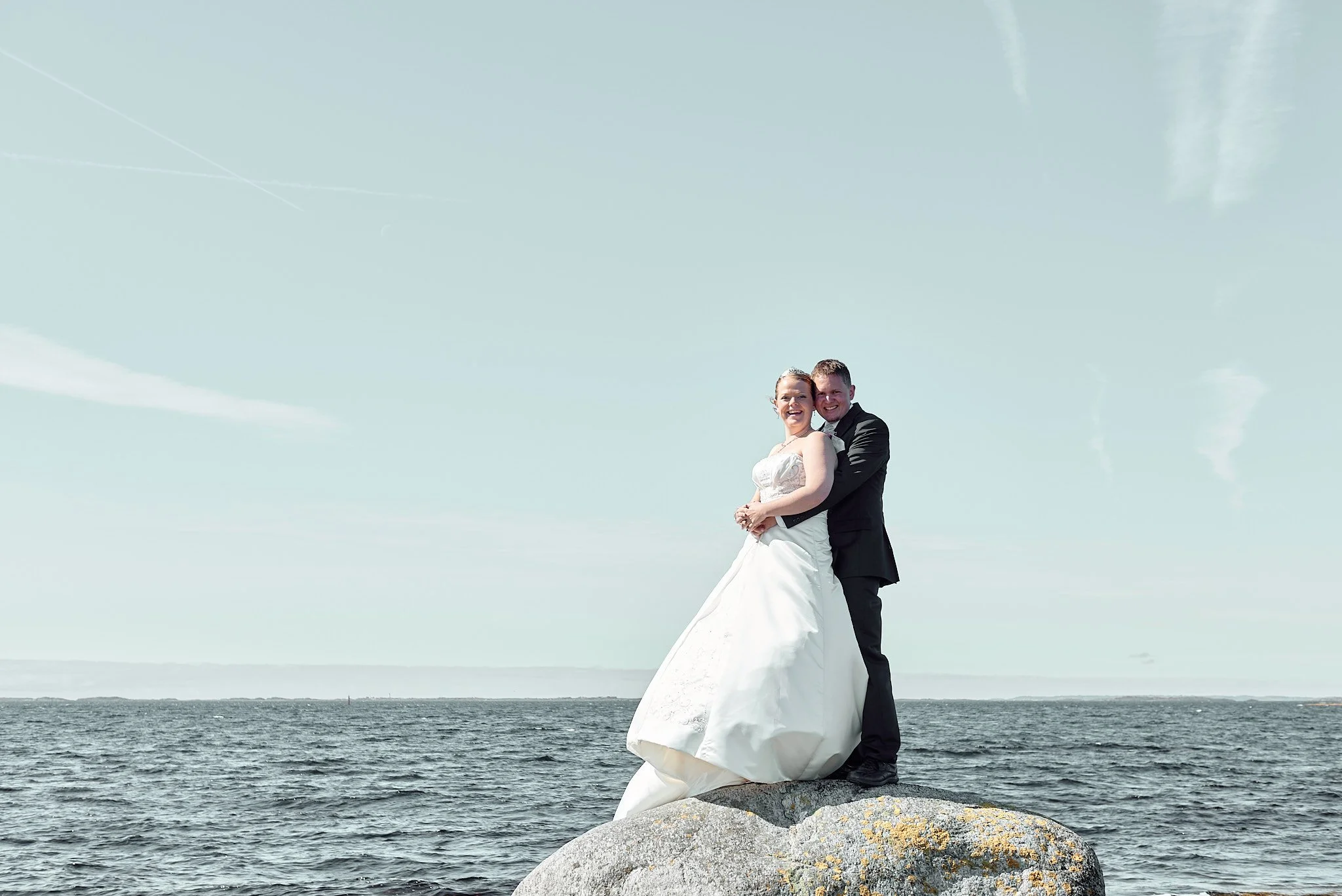 Et brudepar står på en stor stein i havet, med en lys blå himmel og spredte skyer i bakgrunnen.