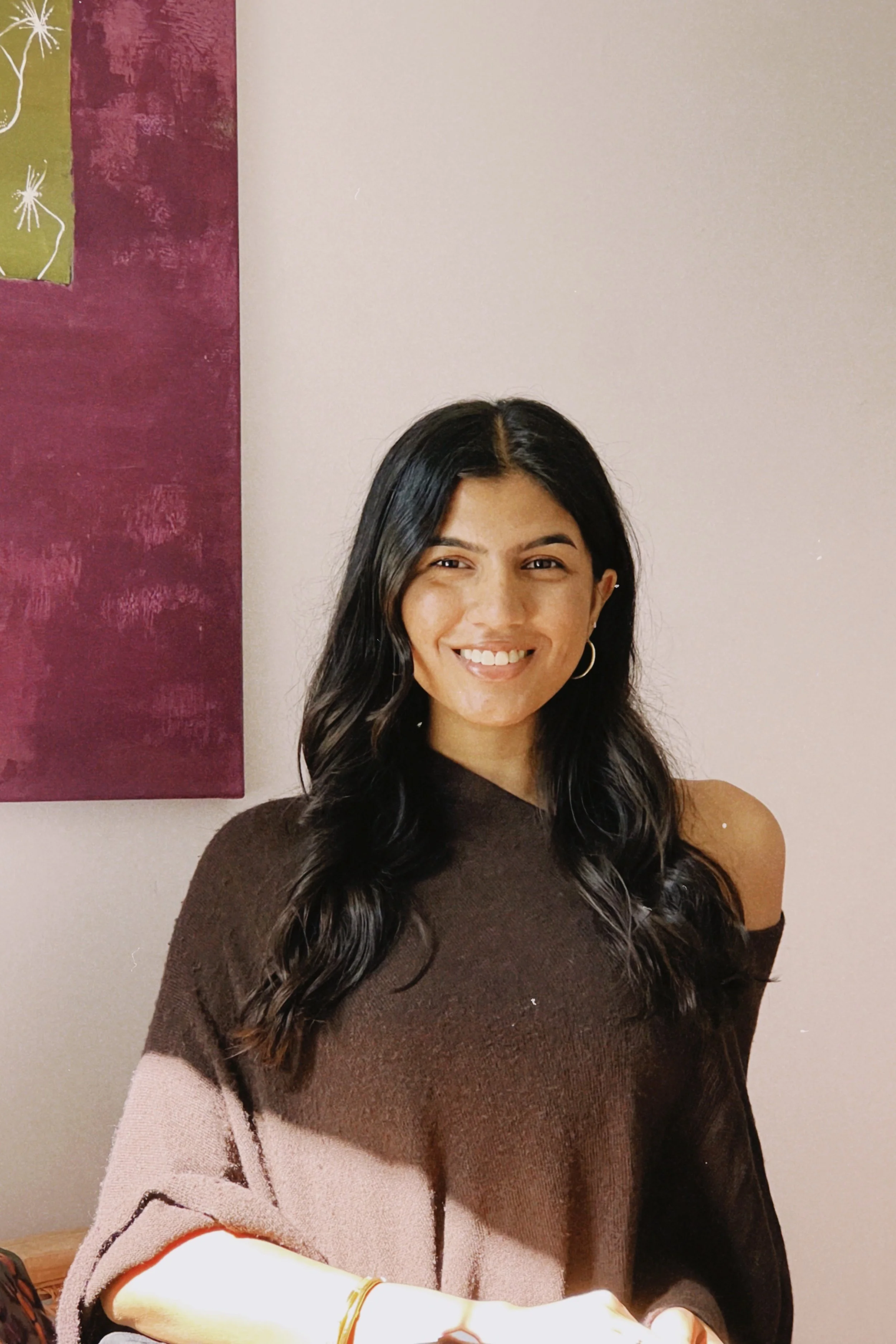 A woman with long dark wavy hair, wearing a brown and pink off-the-shoulder sweater, smiling at the camera, with shoulder-length hair, hoop earrings, sitting against a wall with a maroon and pink painting.