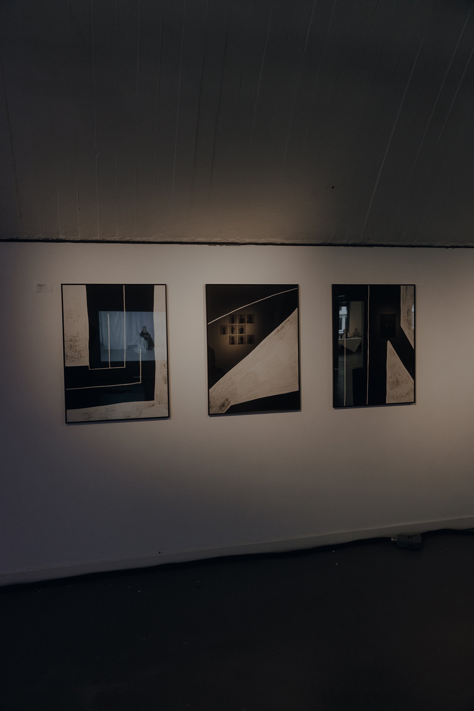 Three framed abstract art pieces hanging on a white wall in a gallery space, with faint lighting and reflections visible on the glass.