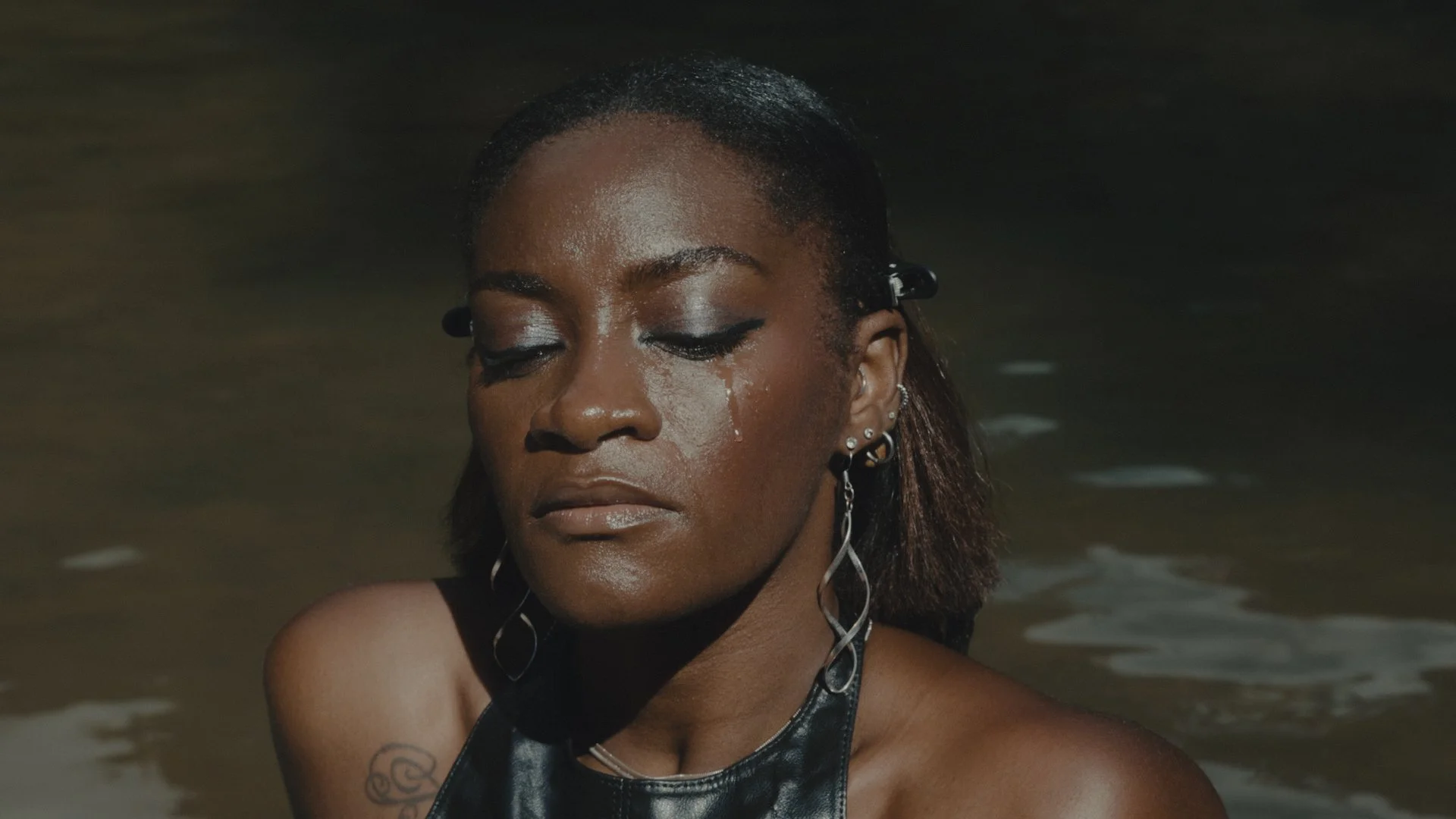 Close-up of a woman with dark skin and wet hair, wearing multiple earrings and a black leather top, standing in water with a calm expression.