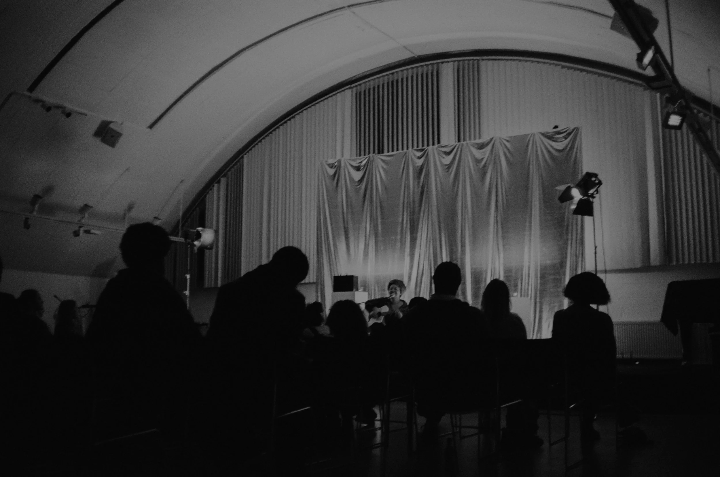 A black and white photo of an indoor stage area with people seated in front, stage lighting, and curtains on the back wall.