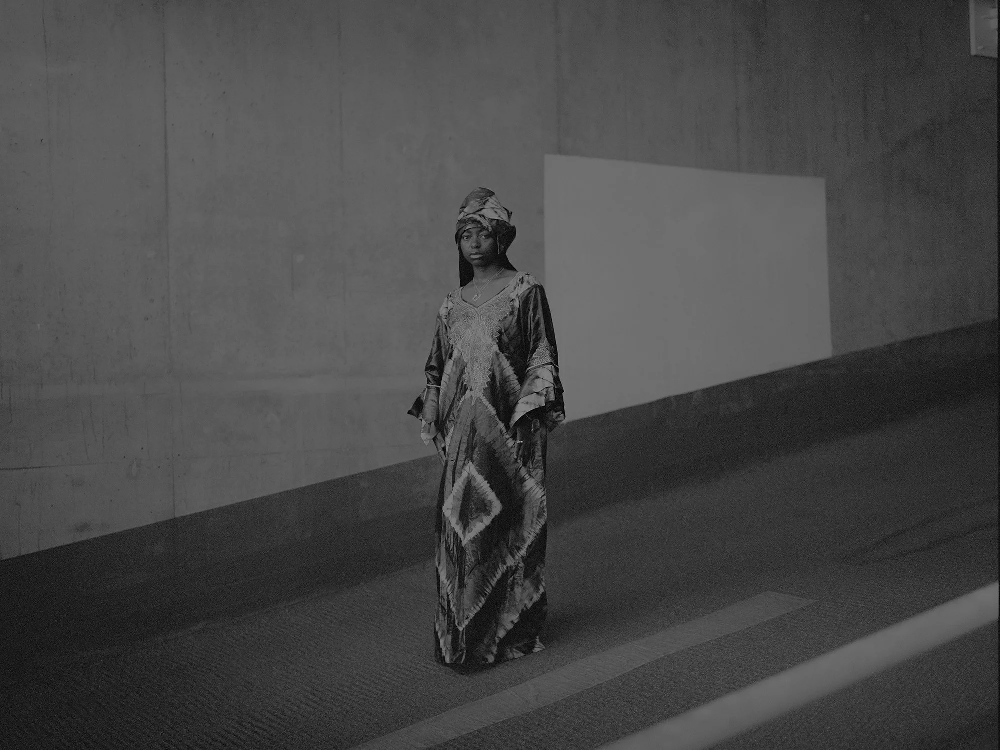 A woman wearing traditional African attire with a headwrap, standing in an indoor gallery or exhibit space with plain walls.