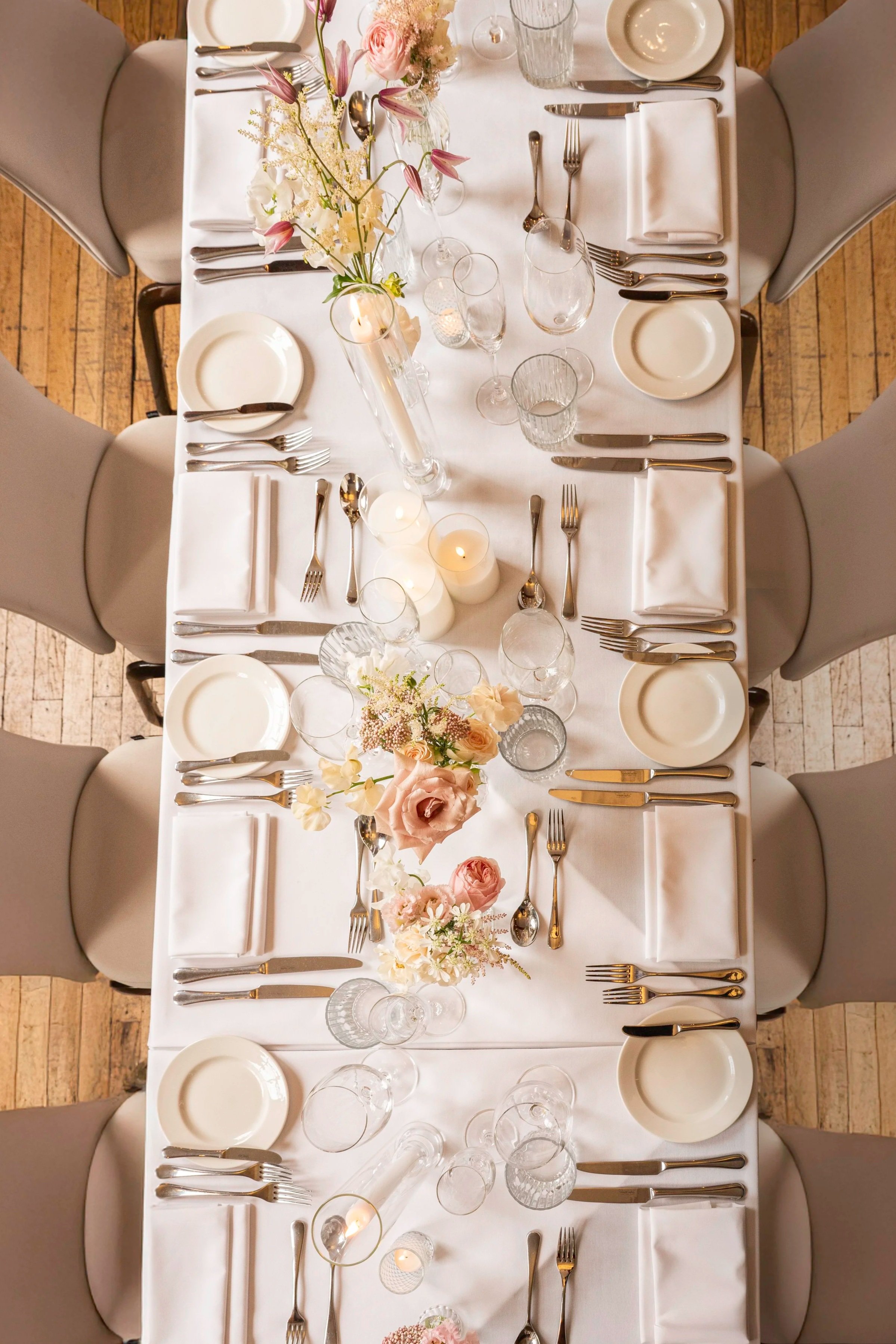 A top-down view of a formal dining table decorated with white tablecloth, floral centerpieces, candles, and set with plates, silverware, and glassware for eight guests.